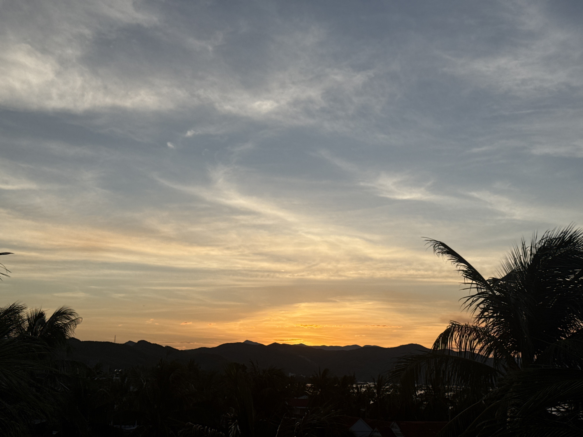 This image captures a serene sunset scene in the city of Song Cau, Vietnam. The sky is a canvas of soft hues, transitioning from a pale blue at the top to vibrant oranges and yellows closer to the horizon. Wispy clouds streak across the sky, catching the fading light. In the foreground, the dark silhouettes of palm trees frame the view, their fronds gently swaying. Beyond them, a range of low, dark mountains forms a dramatic silhouette against the colorful sky. The image conveys a sense of tranquility and natural beauty.
