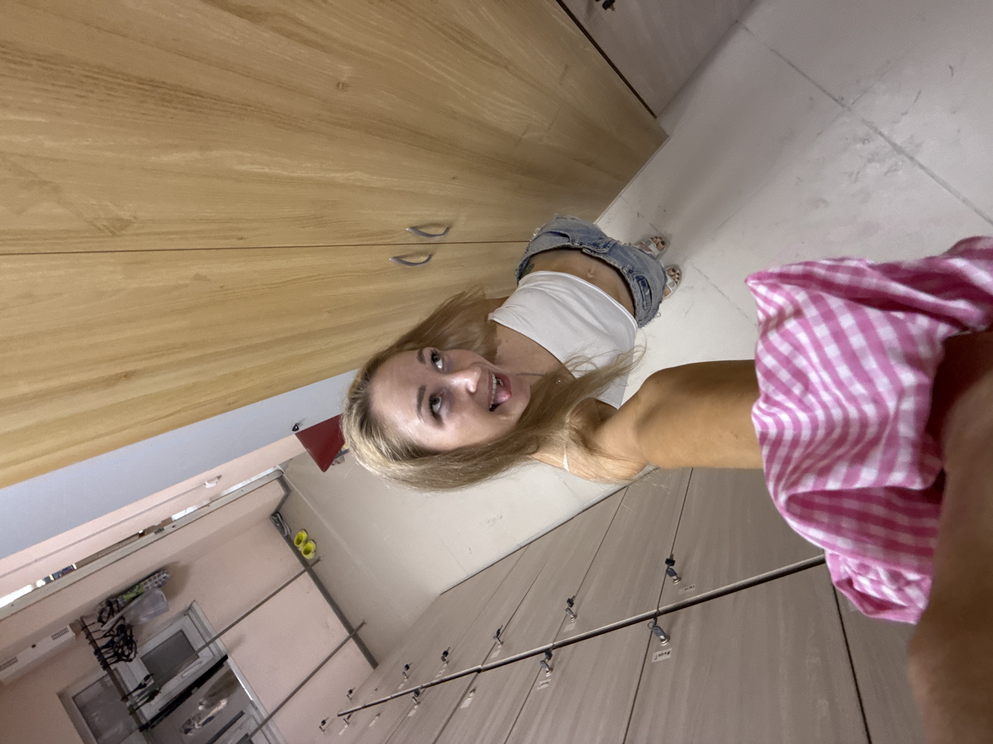 A woman with blonde hair stands in what appears to be a locker room. She is wearing a white top and denim shorts. A pink and white checkered garment is visible in the foreground. The room contains lockers and what seems to be changing area. The location is Chaikovsky, Russia.
