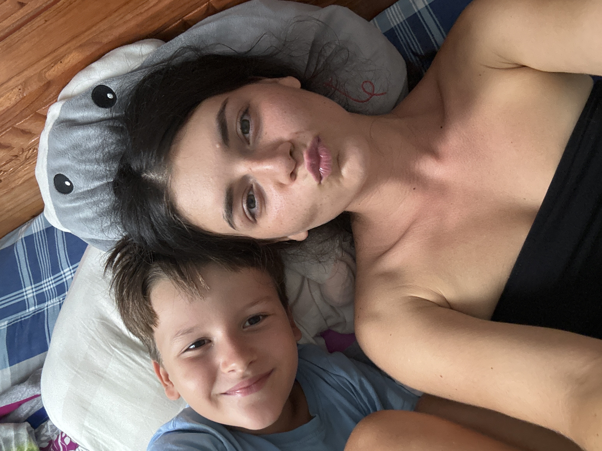 Two people, a woman and a young boy, are lying down in bed. The woman is wearing a black top and has dark hair. The boy is wearing a blue t-shirt and has brown hair. They are both looking at the camera, and the woman is making a pouty face. The setting appears to be a bedroom, with wooden floorboards visible in the background and a blue and white plaid blanket. A grey shark-shaped pillow is also present. The image is taken from a high angle, suggesting a selfie. The lighting is natural, indicating it is daytime. There is no visible text in the image.