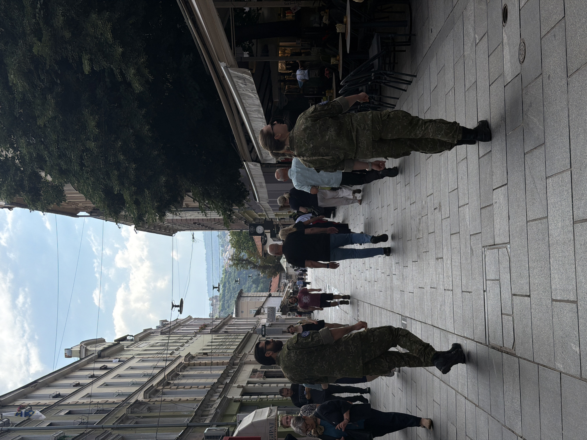 The image depicts a street scene in Sarajevo, Bosnia and Herzegovina, specifically in the Baščaršija district, a historic old town. In the foreground, two individuals in camouflage military uniforms are walking away from the camera on a cobblestone street lined with buildings and outdoor seating for cafes. The man on the left is wearing a green and brown camouflage uniform with a black belt and black boots. He is looking slightly over his left shoulder. The woman on the right is also in a camouflage uniform with her hair in a braid. She is looking straight ahead.
Several other pedestrians are visible further down the street, also walking in various directions. The buildings on either side of the street are tall and have a European architectural style, with multiple stories and windows. On the right side, there are awnings extending from cafes and shops, with tables and chairs set up outside. The sky above is partly cloudy. In the background, a hilly landscape with buildings can be seen. The overall atmosphere appears to be a busy daytime scene in an urban environment.