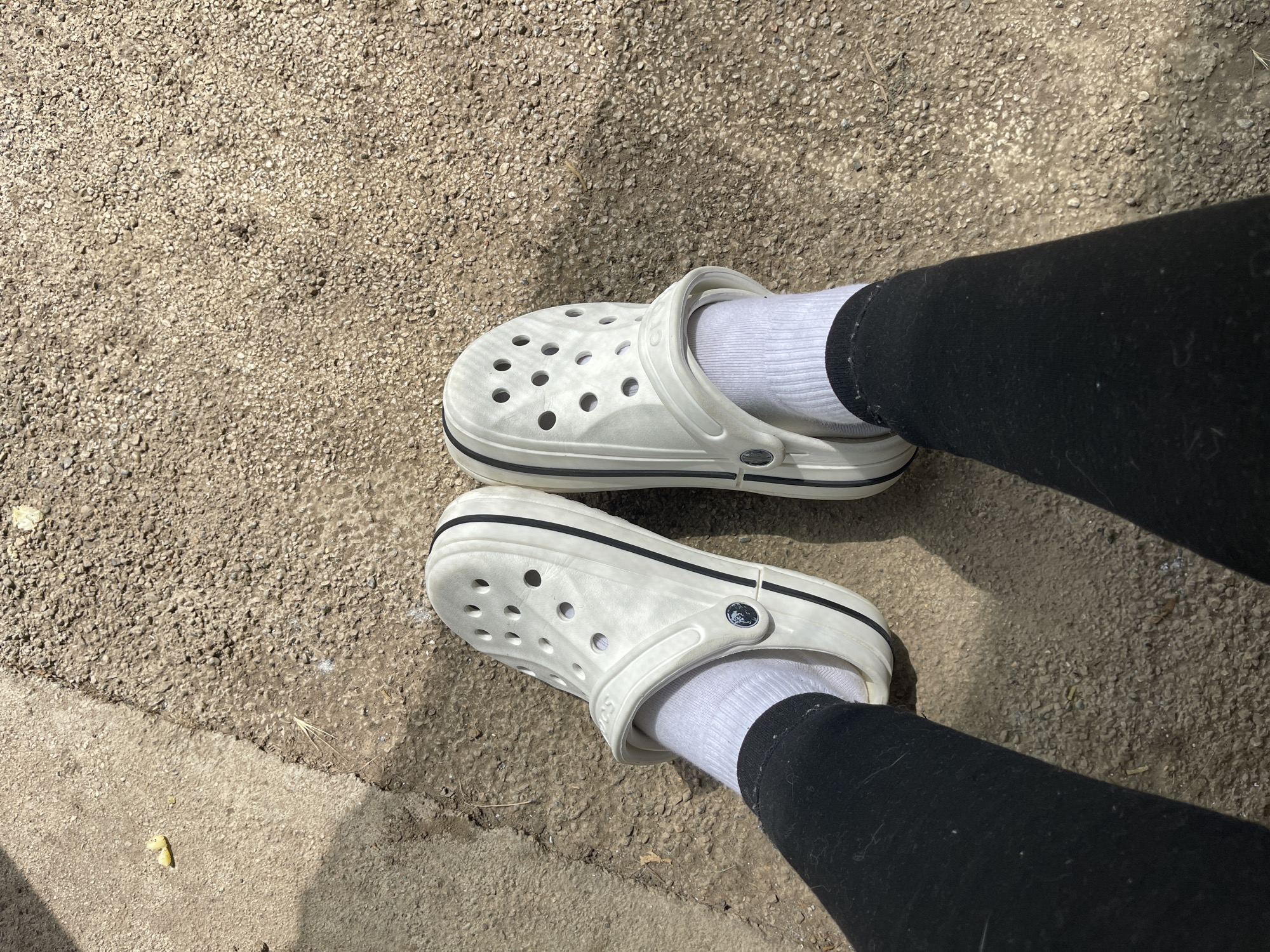 The image shows a close-up view of a person's feet wearing white Crocs with black trim and white socks. The Crocs are positioned on a textured, gravelly surface, likely a sidewalk or pathway. The person is wearing black leggings. The lighting suggests it is daytime, with shadows cast on the ground, indicating the sun is out. The overall scene is casual and appears to be an outdoor setting. The texture of the ground and the presence of small debris like twigs and leaves are visible. There is no discernible text in the image.