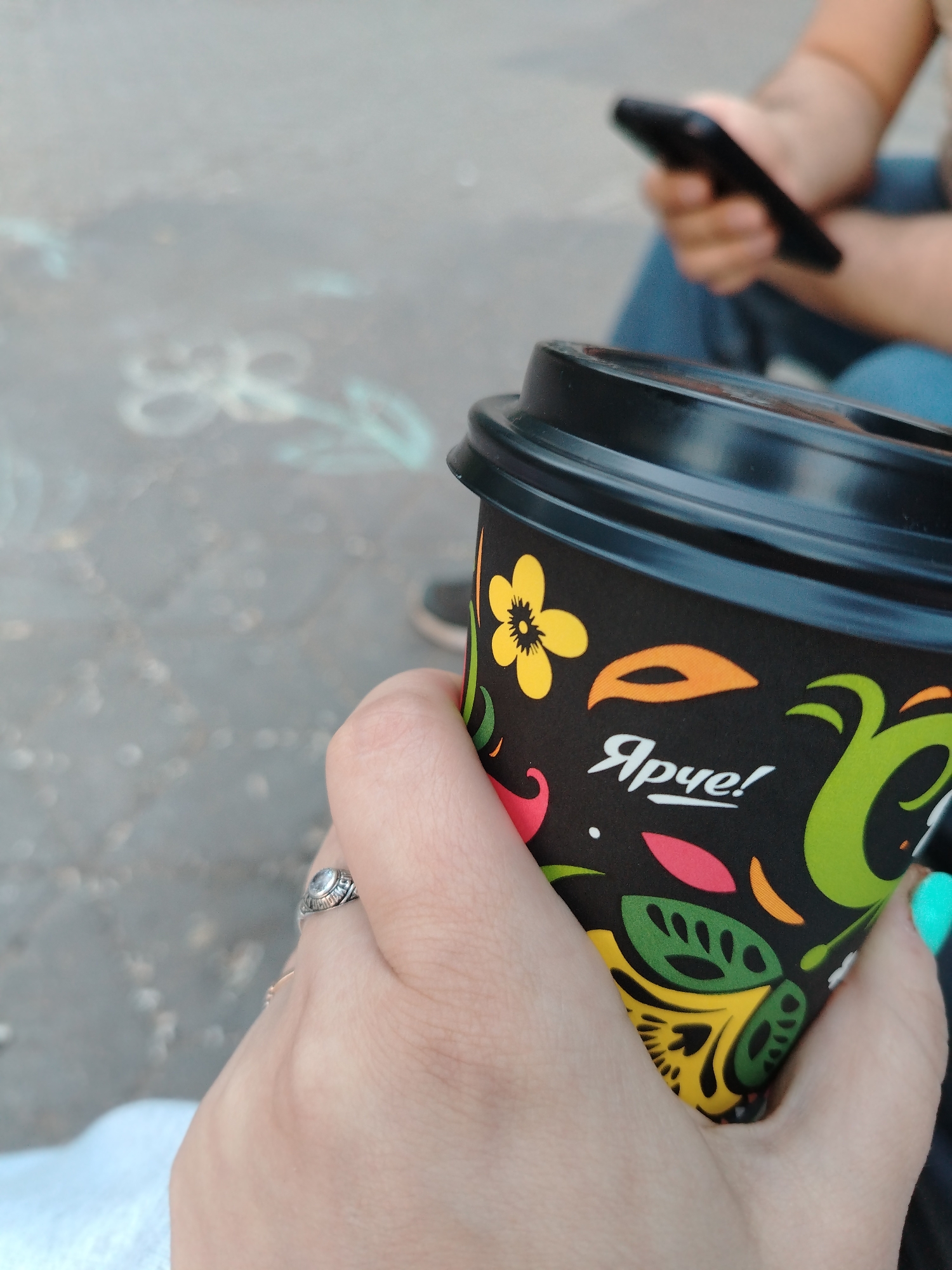 The image presents a close-up, first-person perspective of a human hand holding a black disposable coffee cup. The cup is decorated with a vibrant, stylized pattern featuring yellow, green, orange, and red floral and leaf motifs on a dark background. White Cyrillic text, reading "Ярче!" (Yarche!), is visible on the cup, translating to "Brighter!" or "More vivid!". The hand, appearing light-skinned, wears a silver-toned ring with a clear or light-colored stone on the thumb. The visible fingernail on the little finger is painted a bright turquoise color. In the soft-focus background, another individual is partially visible, holding a black smartphone with both hands. This person is wearing blue jeans. The ground beneath them is a dark, likely paved surface, displaying faint, light-colored markings or graffiti, one of which resembles a stylized "88". The scene is an outdoor urban setting in Yaroslavl, Russia.