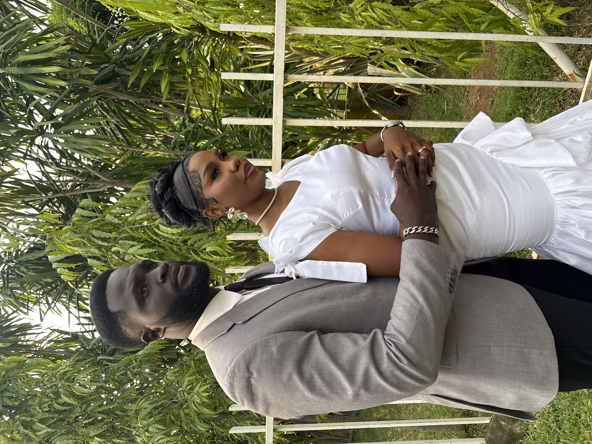 This image depicts a couple posing outdoors, likely for a wedding or engagement photoshoot, in Barikin Gidar Kanat, Nigeria. The woman is wearing a white dress with a pearl necklace and elaborate earrings. Her hair is styled in braids, and she has dramatic eye makeup. The man is dressed in a grey suit with a white shirt and black tie. They are holding hands, with the woman's left hand resting on the man's arm and his hand gently holding her abdomen. A ring is visible on the woman's left hand. The background consists of lush green foliage, suggesting a garden or natural setting. A white metal fence is also visible, partially obscured by the greenery. The lighting indicates it is daytime, likely with bright sunshine. The couple's poses and gazes suggest a romantic and celebratory mood.