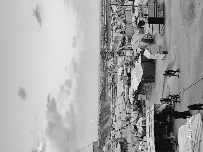 This black and white image presents a wide, aerial-like view of a densely packed refugee or displacement camp. The scene is filled with numerous makeshift structures, primarily tents and shelters fashioned from tarpaulins and various materials, extending across a barren, undulating landscape under a partly cloudy sky.
An unpaved road or path traverses the camp, serving as a thoroughfare for several individuals seen walking or standing. In the mid-ground, a small group of people moves along the path, while other figures are visible near the rudimentary dwellings. The structures appear tightly clustered, suggesting crowded living conditions. Some of these structures resemble small market stalls or shops, with one clearly displaying a sign.
The lighting indicates daytime, likely mid-day or afternoon, casting soft shadows. The distant horizon reveals more of the camp and arid hills, with a solitary communication tower visible. The monochromatic tone of the photograph emphasizes the stark reality and challenging environment of the camp.
Visible text includes Arabic script on a banner or sign on one of the structures, which reads "المجد" (Al-Majd - The Glory/Majesty) followed by "للأجهزة" (for appliances/devices), indicating a small store. A "Galaxy A14" watermark is present in the bottom right corner of the image.