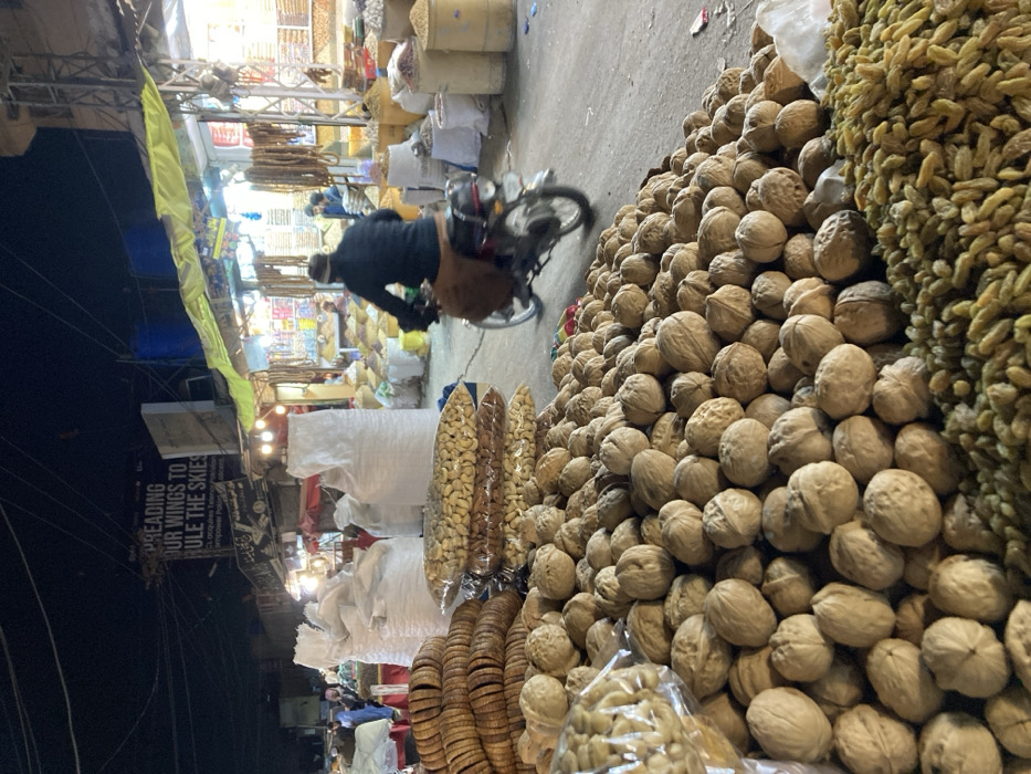 A vibrant marketplace presents a medley of nuts and dried fruits. Walnuts dominate the foreground, piled high. Cashews, figs, and raisins also feature prominently. A man on a motorcycle weaves through the scene, navigating past white sacks potentially containing more goods. Illuminated storefronts and overhead lights hint at an evening setting, with buildings and power lines visible in the background.
