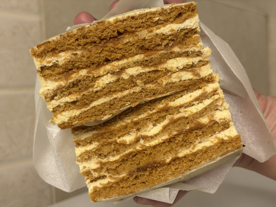 Two layered honey cakes are held together in one hand, wrapped in a sheet of parchment paper. The cakes are rectangular with multiple thin layers of golden brown cake alternating with layers of light yellow cream. The layers are slightly uneven, adding to the rustic appearance. The top layer of cake is also frosted with a light yellow cream. The cakes are slightly offset, with the top piece angled slightly upward.