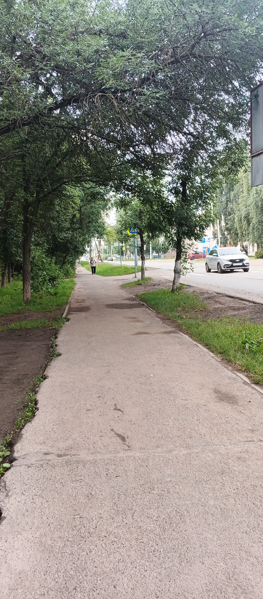 The image depicts a sidewalk and a street in a city. On the left side of the image, there is a sidewalk lined with trees and green grass. A person, appearing to be a woman in a dark jacket and light-colored pants, is walking away from the camera on the sidewalk. On the right side of the image, there is a street with a white car driving on it. The street is also lined with trees. In the background, there are buildings and other cars. The sky is overcast. The scene appears to be on a street in Izhevsk, Russia, possibly in a residential area with a commercial or traffic area nearby.
