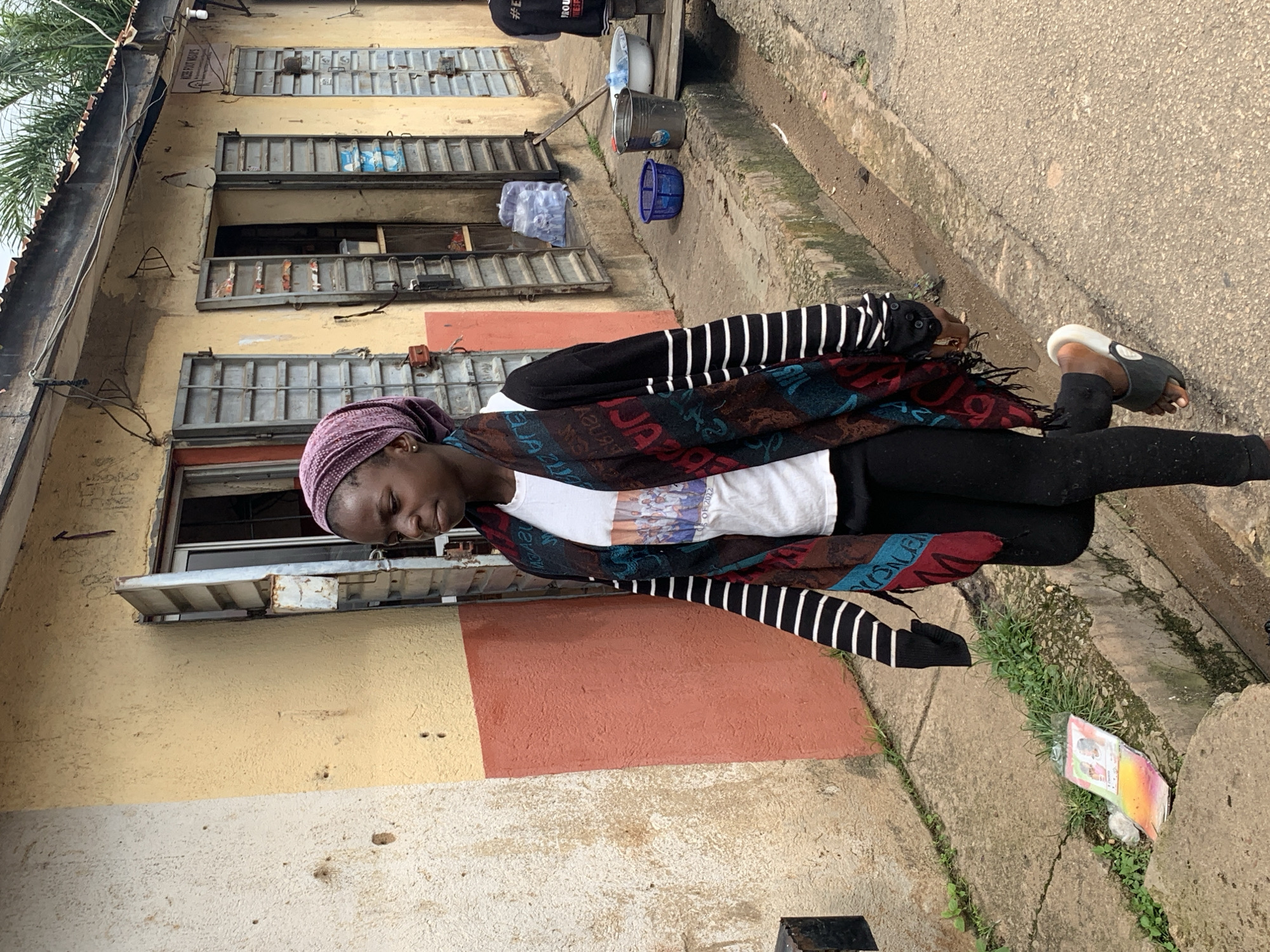A young Black girl, wearing a purple headscarf, a black cardigan over a white t-shirt, black leggings, and flip-flops, walks along a street in Jos, Nigeria. She is seen from the chest down, walking with her head turned away from the camera. The street is paved with asphalt, with a concrete gutter on the left. A yellow and pink building with metal-shuttered windows and an open doorway stands behind her. Several plastic bottles of water and two buckets are visible on the sidewalk. The building appears to be a shop. A piece of colorful paper with images is on the ground near the gutter. The sky is overcast, suggesting it might be a cloudy day.