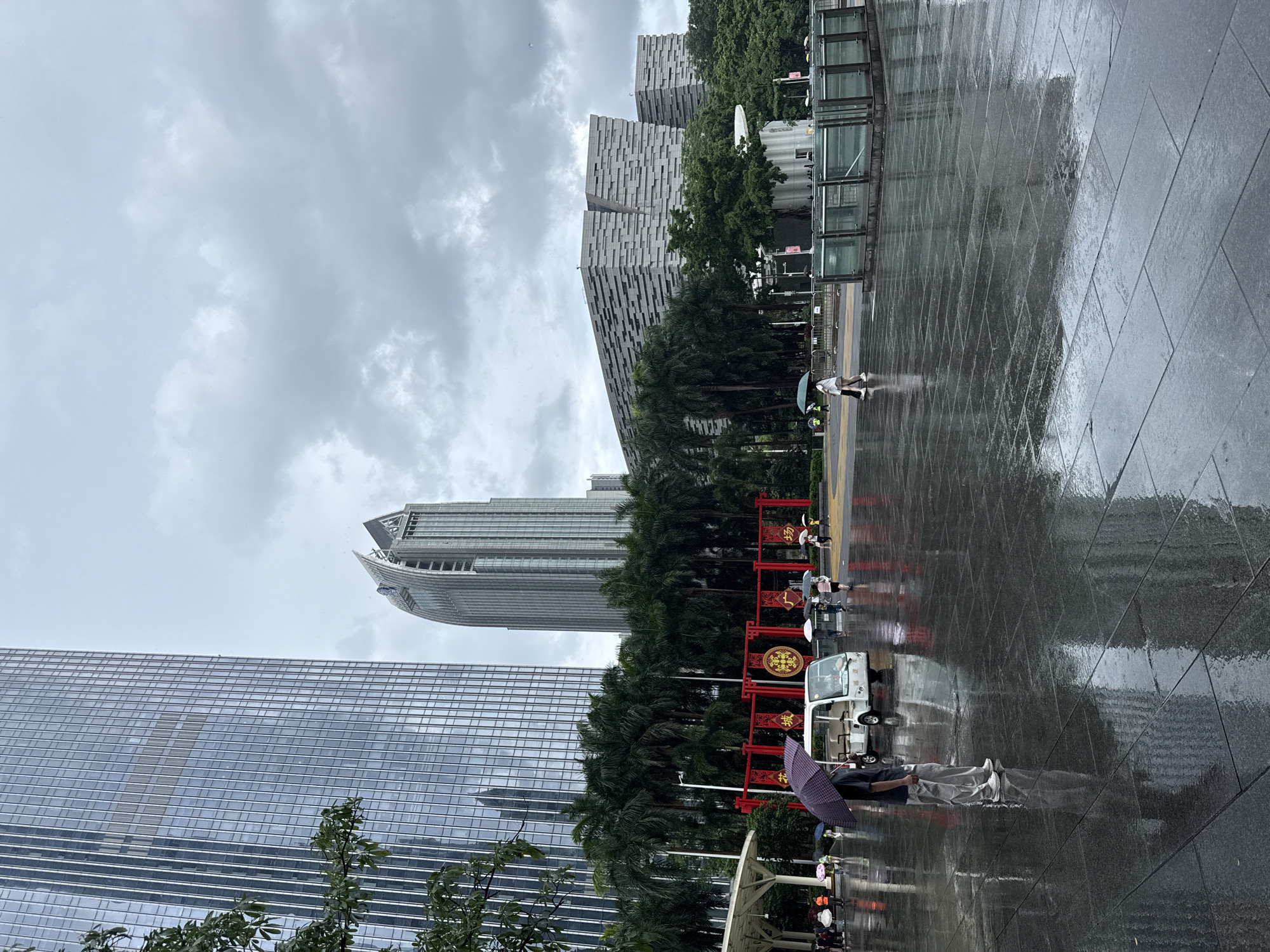 The image depicts a wet, overcast day in Tianhe District, China. The ground, paved with large, dark tiles, is saturated with rain, reflecting the surrounding architecture and sky. In the foreground, a person stands under a dark umbrella, looking to their left. A small white electric vehicle is parked behind them.
Dominating the background are several modern skyscrapers. The tallest, on the left, is a sleek glass-fronted building. To its right, another skyscraper features a distinctive, curved, pointed roofline. Further back and to the right, a long, low building with a textured, stone-like facade is visible.
Between the buildings, a row of palm trees adds a tropical element. A red archway with golden Chinese characters stands prominently, adorned with circular decorations. Several other people, many holding umbrellas, are scattered across the plaza, moving in different directions. The sky is filled with heavy, grey clouds, indicating ongoing or recent rainfall. The overall atmosphere is one of a bustling urban environment experiencing inclement weather.