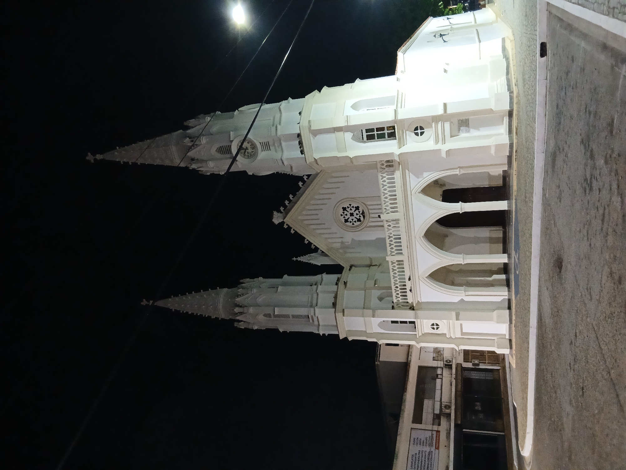 The image shows the Iglesia de San Juan Evangelista, a white-painted church with two towers, located in Juan Griego, Venezuela. The church is illuminated by streetlights at night, and the sky is dark. Part of a building is visible in front of the church. The edge of a street is visible on the right. A sign on the building reads "LAVAPLATOS DESINPECTANTES DESENGRASANTE CLORO COCLON TES-CERA ANTE CHAMPU".