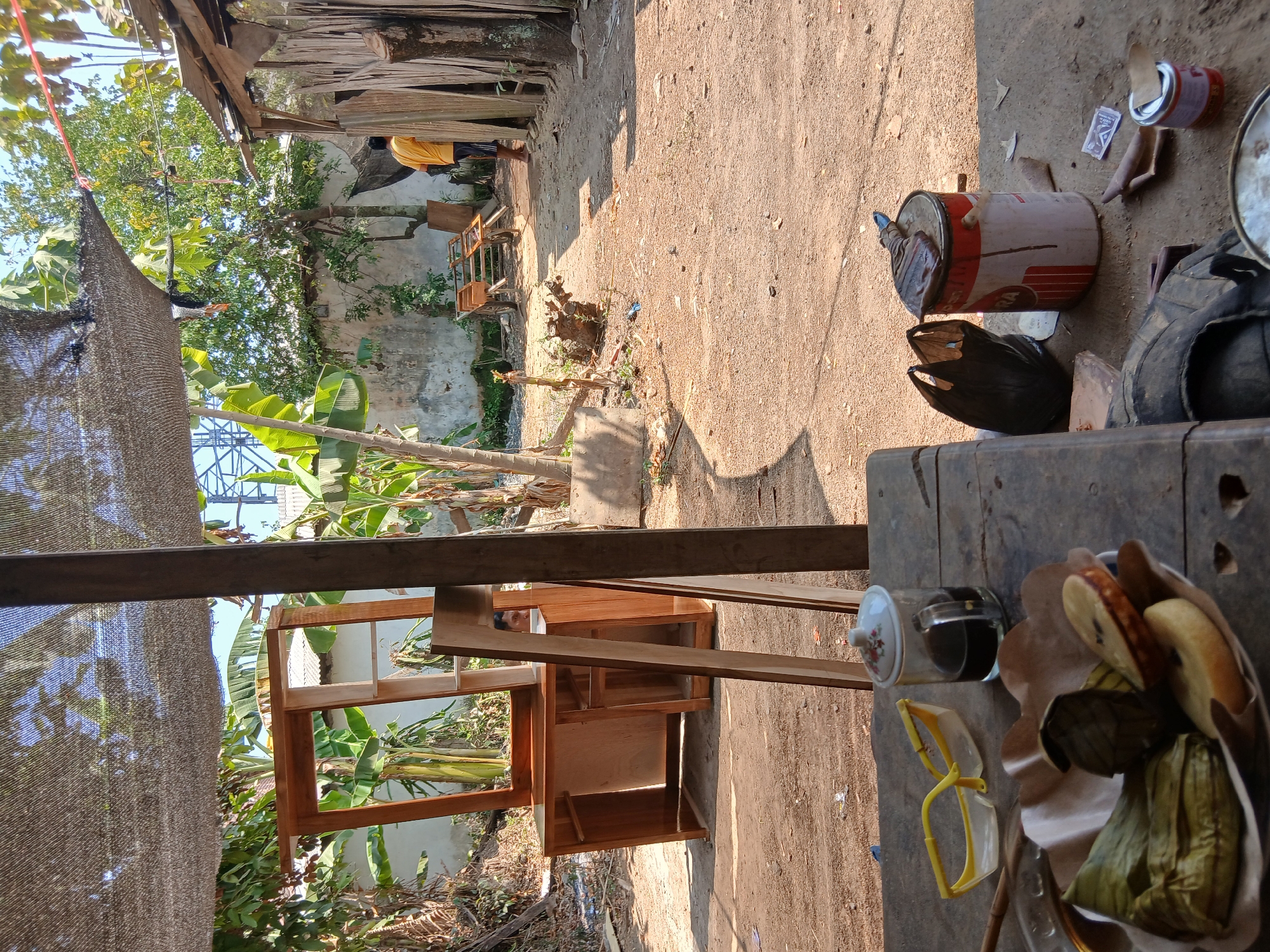 In Tenggarang, Indonesia, this image depicts an outdoor workshop area, likely a furniture-making or repair shop, under a woven sunshade. A partially assembled wooden furniture piece, possibly a cabinet or desk, stands prominently on the left. In the foreground, a rustic wooden table holds a glass of dark liquid, yellow safety glasses, and a plate of food wrapped in banana leaves, along with some pastries. To the right, a large metal paint can with a brush on top sits on the ground, next to a black plastic bag and a smaller can of paint. In the background, a man wearing a yellow t-shirt and dark shorts walks away from the camera. The ground is covered in dirt and small debris, with banana trees and other vegetation visible around the workshop. A dilapidated wall and wooden structures suggest a rural or semi-industrial setting.