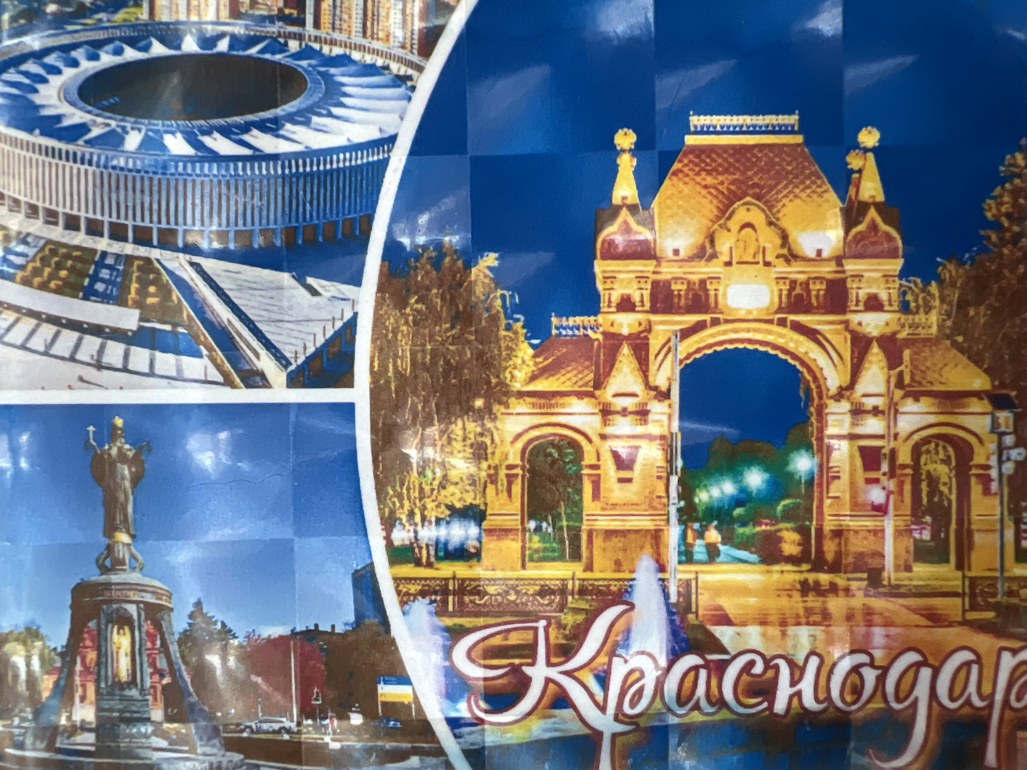 The image depicts a collage featuring several landmarks in Krasnodar, Russia. The top left shows Krasnodar Stadium, a circular structure with a visible inner ring and supporting pillars. Below it is a statue of Catherine the Great, standing atop a pedestal with other figures and architectural elements. The main focus is a golden-colored archway or gate with elaborate detailing and surrounding trees, with "Krasnodar" written in white script below it. The background is a blue hue, and there are signs of water or reflections in the foreground.