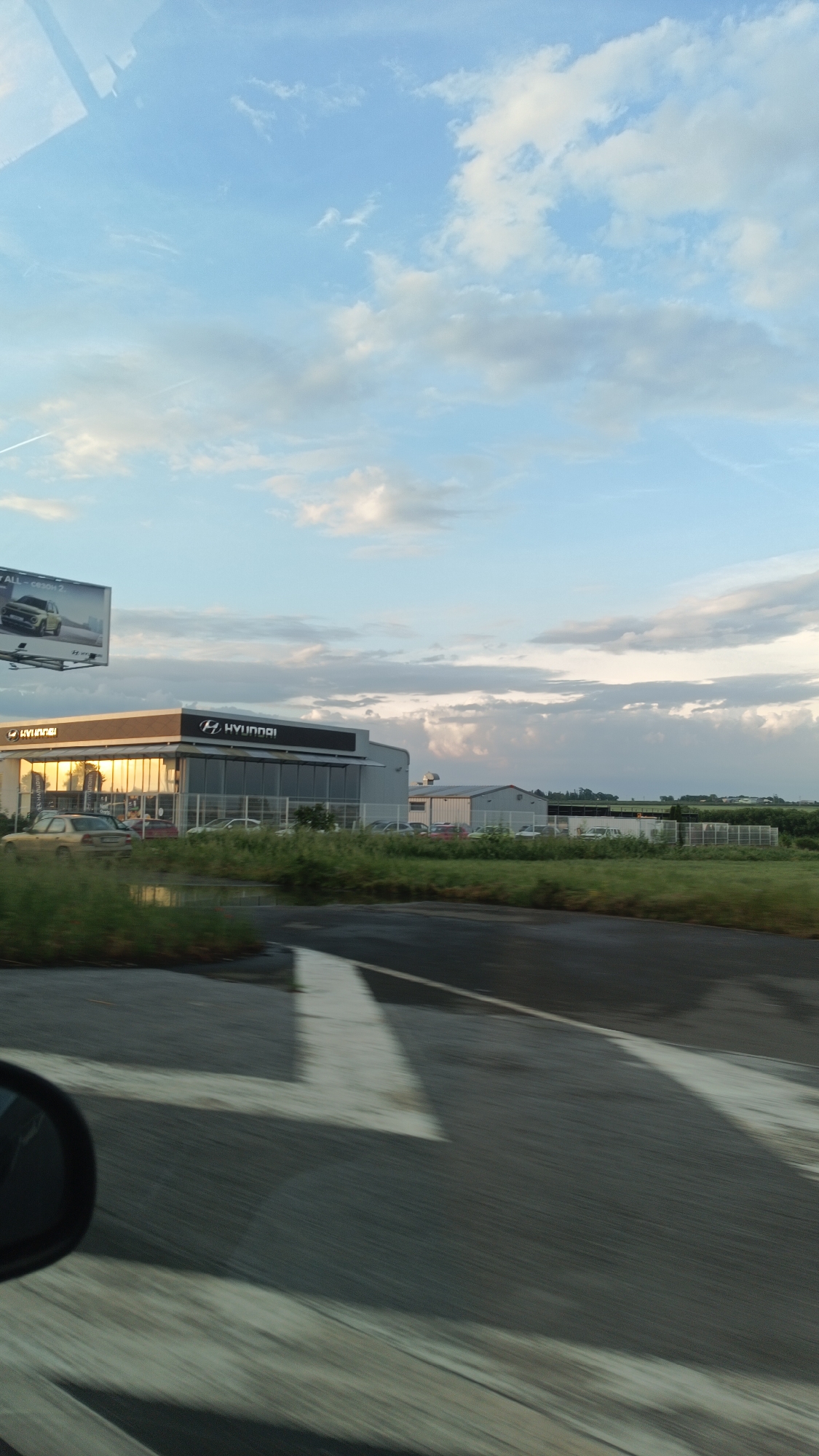 The image shows a Hyundai dealership in Burgas, Bulgaria, as seen from a moving vehicle. The dealership building is visible, with the Hyundai logo prominently displayed. There is a billboard with a car advertisement on the left of the image. The foreground shows the road with painted markings, and the side mirror of the vehicle is visible in the lower-left corner. The sky is partially cloudy. There are fields and other buildings in the background.
