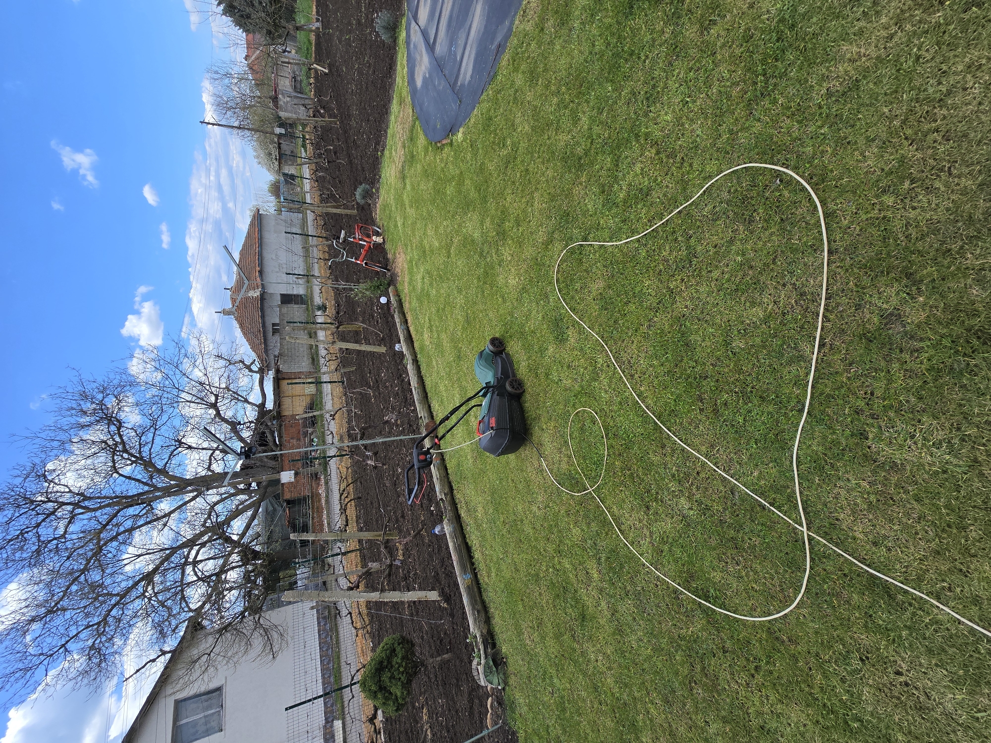 The image shows a garden scene in Stara Reka, Bulgaria. A green lawn is the primary subject, with a dark grey and green lawnmower positioned on it. A long, white electrical cord extends from the lawnmower across the grass. Adjacent to the lawn is a garden plot with exposed dark soil and a fence line. A building is visible in the background, partially obscured by a bare tree. The sky is blue with scattered white clouds.