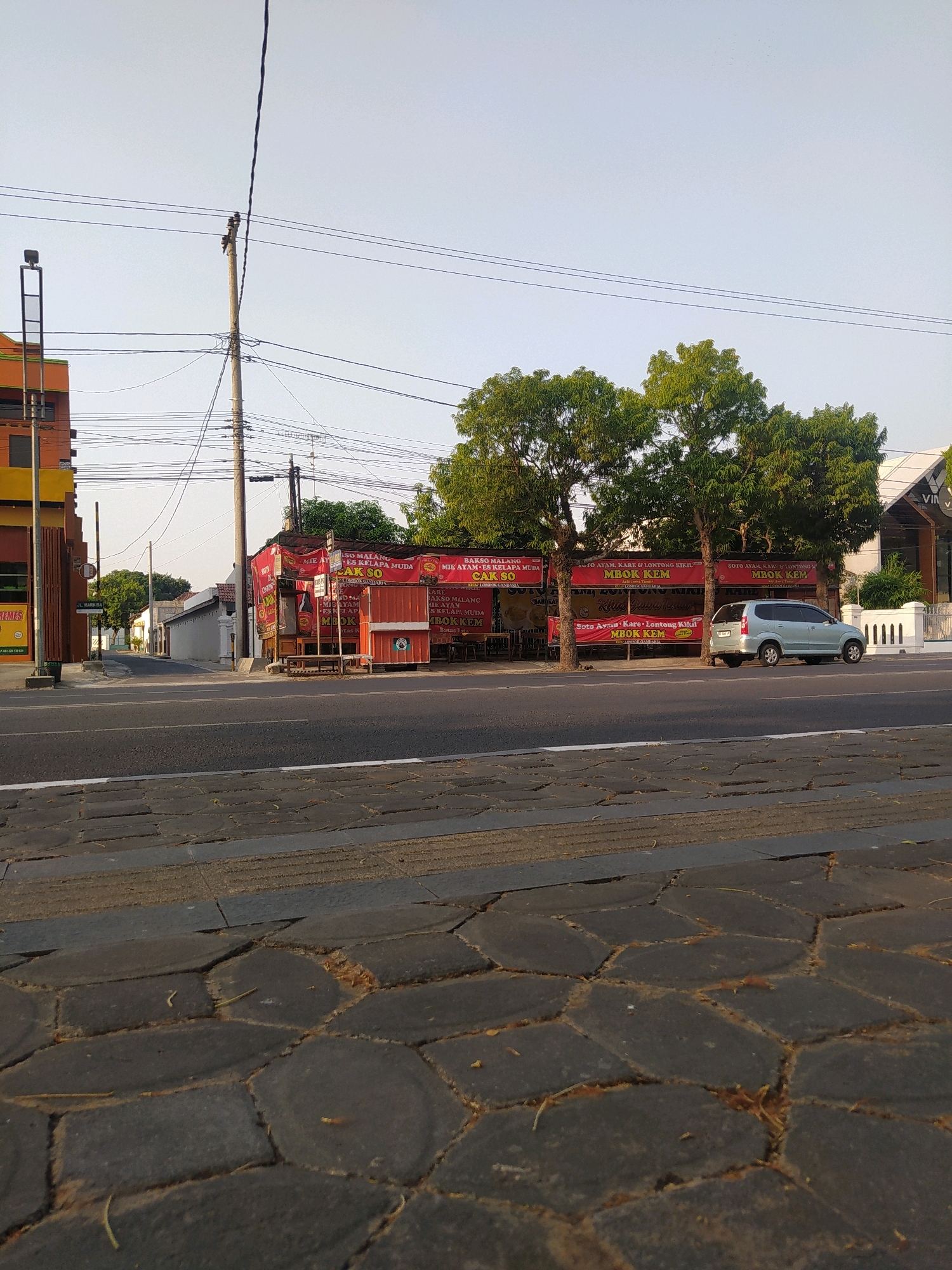 A street scene in Papahan, Indonesia, shows a roadside eatery with red banners displaying food offerings, such as "MIE AYAM KARE & LONTONG KIKIL" and "MBOK KEM". A silver minivan is parked in front of the eatery. The street is paved with dark hexagonal stones, and a sidewalk with similar paving is visible in the foreground. Overhead power lines crisscross the sky, supported by a utility pole. The time of day appears to be late afternoon, indicated by the warm lighting and long shadows. In the background, there is a building with a yellow and orange facade, and several trees with green foliage line the street. The sky is clear and light blue.