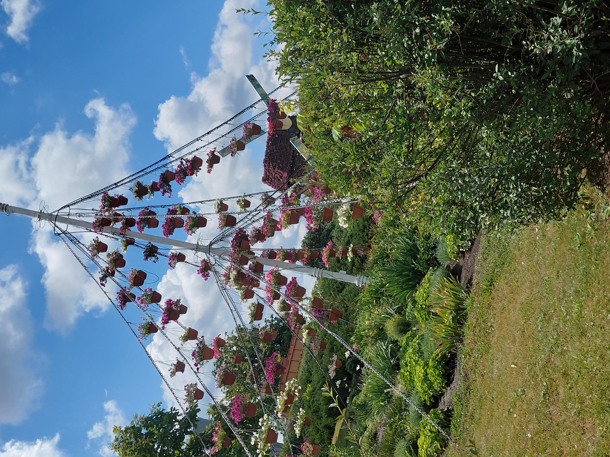 The image shows an outdoor scene in Kovalivka, Ukraine, featuring a large, conical-shaped structure adorned with numerous hanging potted flowers. This structure, possibly a decorative art installation or a unique garden feature, is supported by a central pole and appears to have tiered, angled elements from which the flower pots are suspended by chains. The pots themselves are small and brown, containing pink, white, and possibly purple petunias.
The background consists of a bright blue sky with scattered white clouds. To the left of the central structure, there is a lush green tree. The ground below is covered with various types of vegetation, including a manicured lawn in the foreground, and meticulously planted flower beds and shrubs with various shades of green. In the middle ground, to the right of the hanging flower structure, a large, dense green bush dominates the right side of the frame, partially obscuring the view of the ground and some of the vegetation behind it. A hint of a windmill-like structure covered in red flowers is visible behind and to the right of the hanging flower installation. The scene is well-lit, suggesting it was taken on a sunny day. There are no people visible in the image.