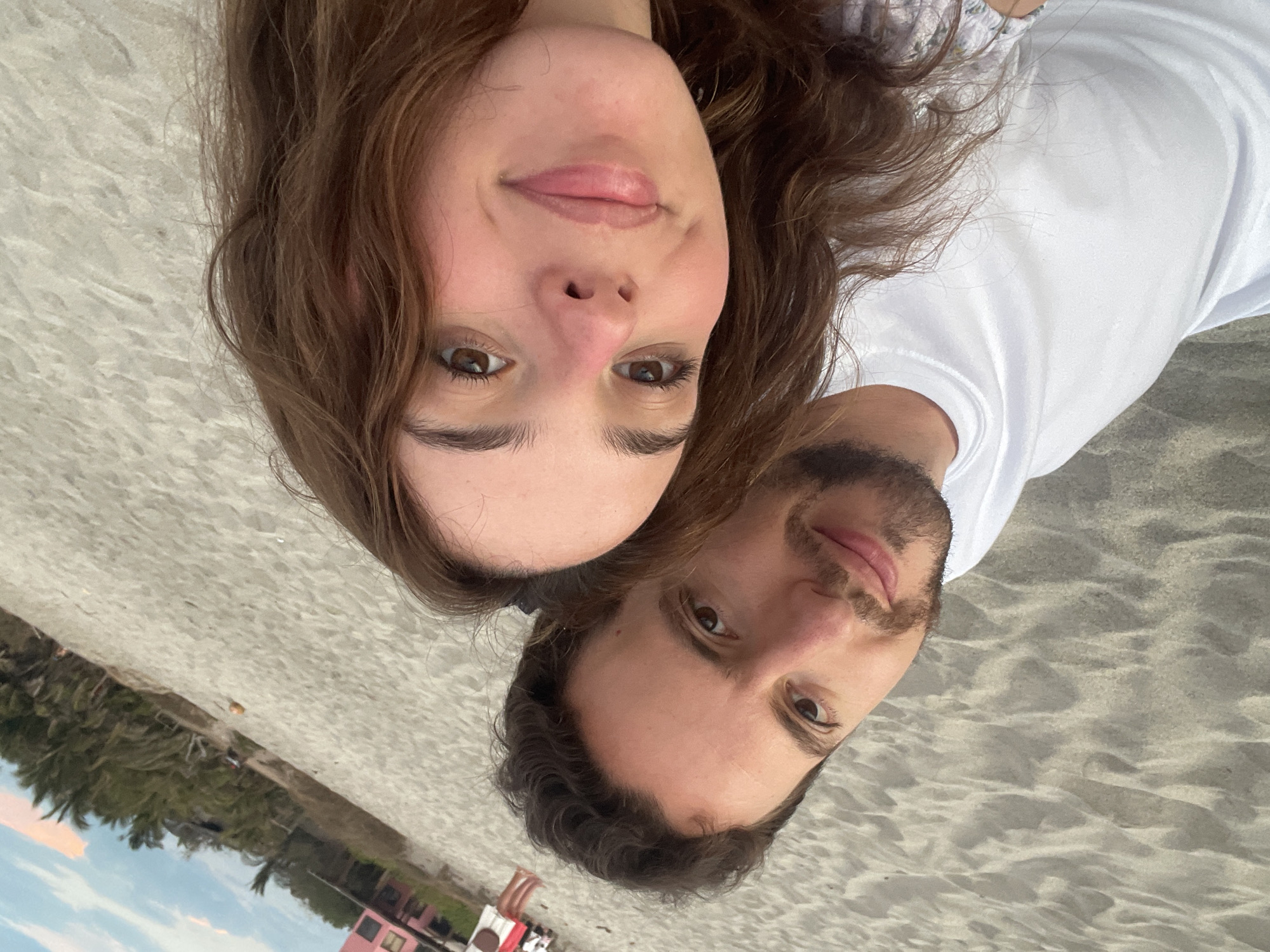 A man and a woman pose for a selfie on the beach in Brisas de Zicatela, Mexico. The woman is wearing a floral shirt and has brown hair. The man is wearing a white t-shirt and has dark hair and a beard. The background shows palm trees, a pink building, and other people on the beach.