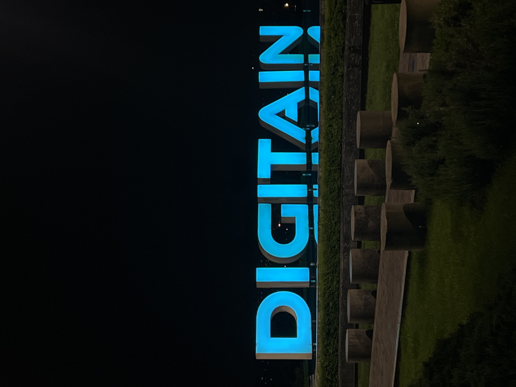 The image shows an illuminated vertical sign that reads "DIGITALAIN" in blue lettering against a dark background. The sign is positioned next to a green space with a row of cylindrical concrete structures or benches. The surrounding environment is dark, suggesting it is nighttime. The location is Unknown, Unknown.
