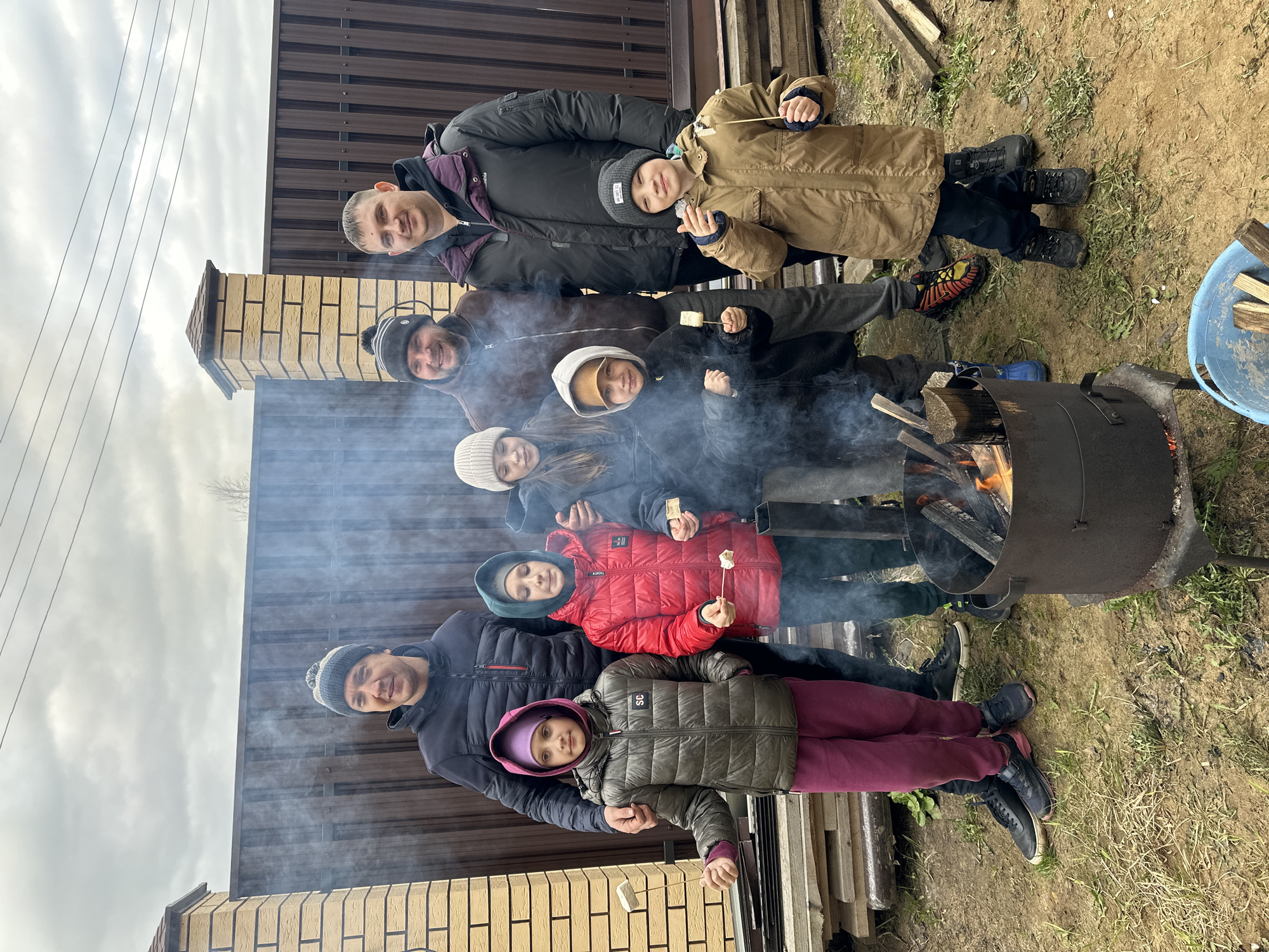 A group of eight people are standing together outdoors, presumably toasting marshmallows over a fire pit. There are two adults at the top of the frame and another at the bottom. In between are five children. Each person is wearing a winter jacket and some form of hat. Three of the children are holding marshmallows on sticks. The fire pit is made from dark metal and flames are burning inside. In the background is a brown fence and a brick wall. The ground is covered in grass and dirt. The sky is overcast. The photo appears to have been taken in слобода Старая, Russia.