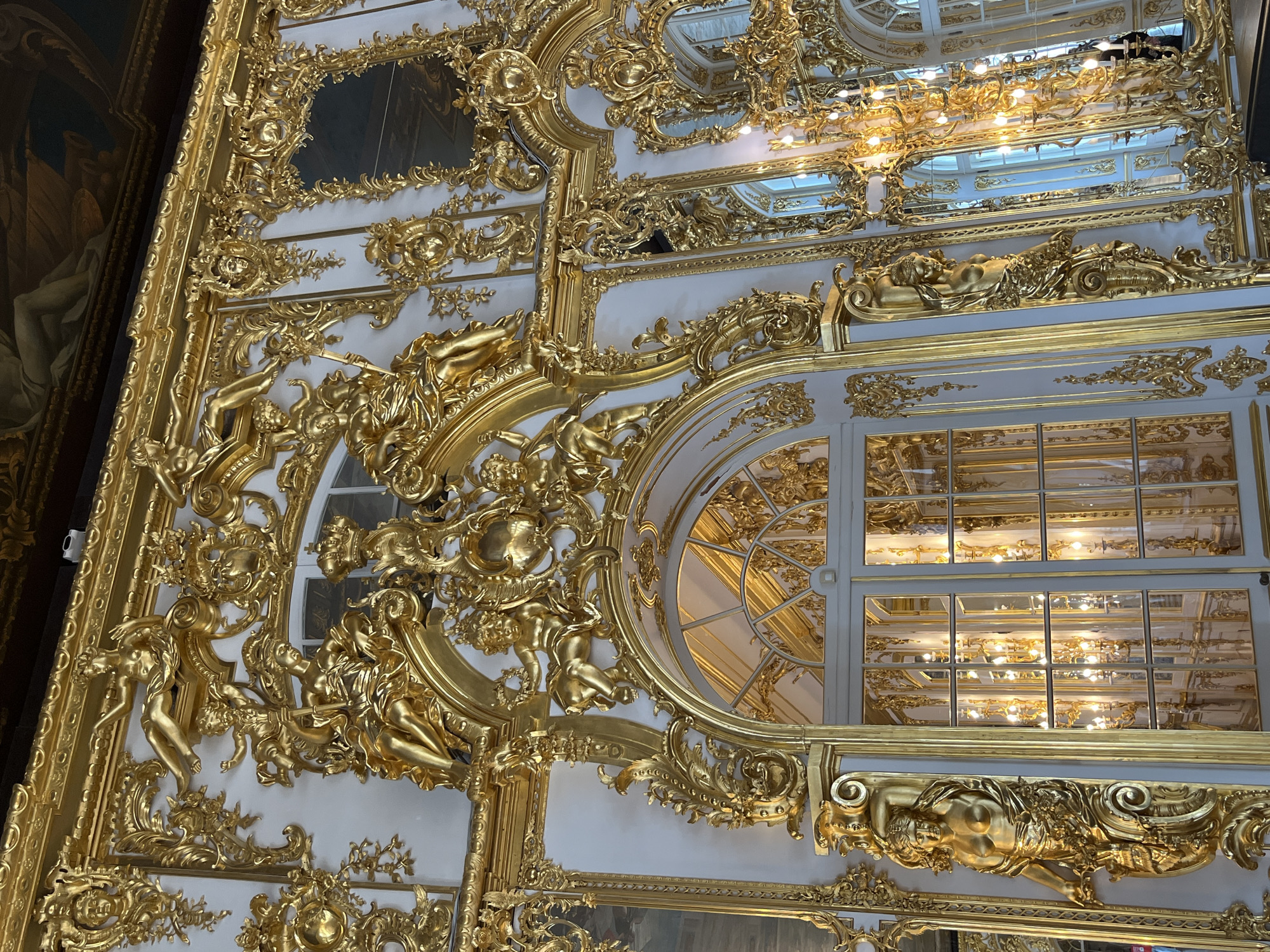 The image shows a section of elaborately decorated interior. The decor is primarily white with gold ornamentation, including sculpted figures, floral patterns, and intricate designs. Windows, some arched, are visible, reflecting interior lighting. The style suggests a Baroque or Rococo influence. The location appears to be an ornate palace or historical building.