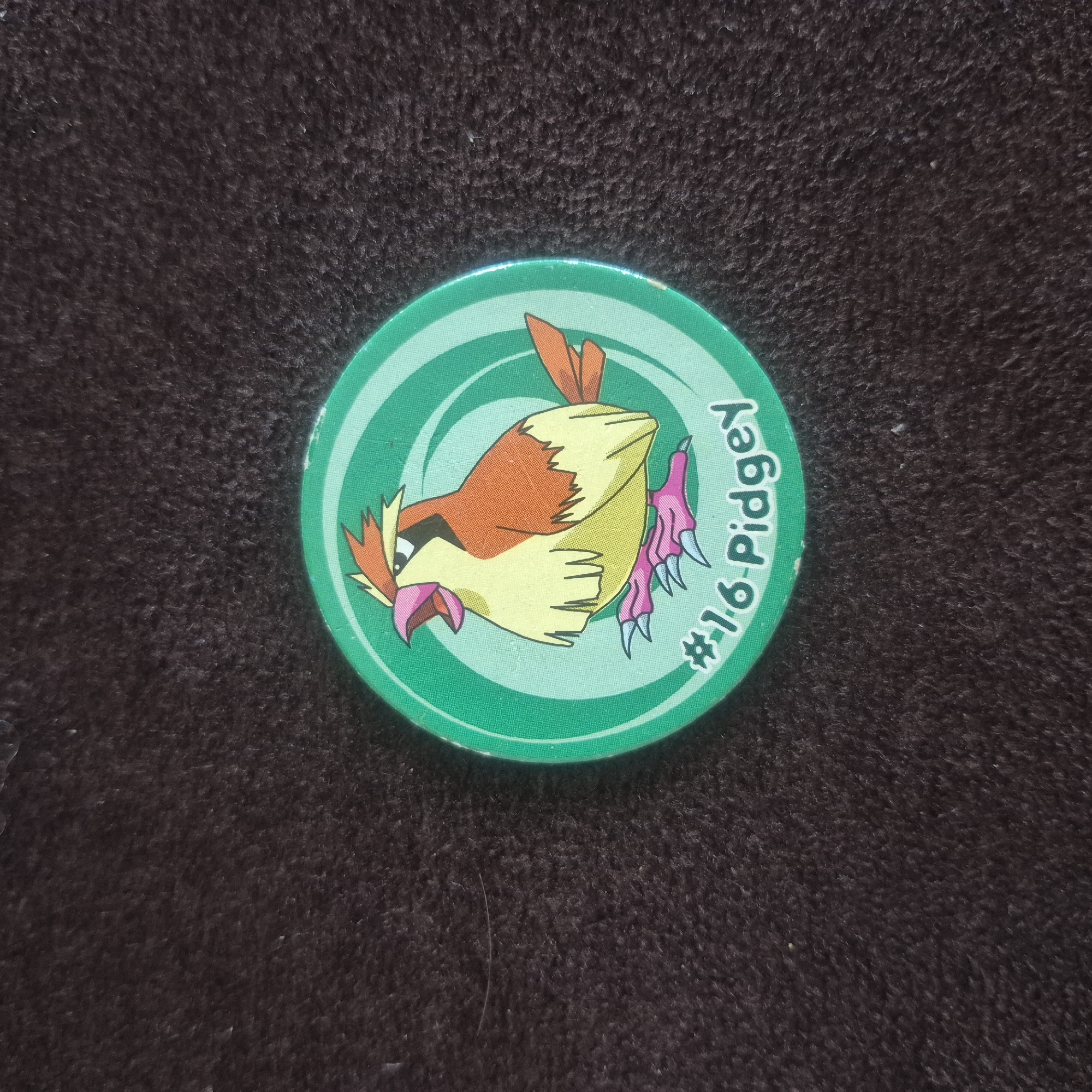 A round, green Pokemon trading disc featuring the character Pidgey is placed on a dark, fuzzy surface. The disc has concentric rings and a drawing of Pidgey in the center. Below the image of Pidgey, the text "#16 Pidgey" is visible. The dark surface appears to be a fabric, possibly a rug or blanket, and shows a textured pattern. There are no people or any other objects visible in the image. The lighting suggests an indoor setting. There is no information within the image to suggest its location in Kępno, Poland.