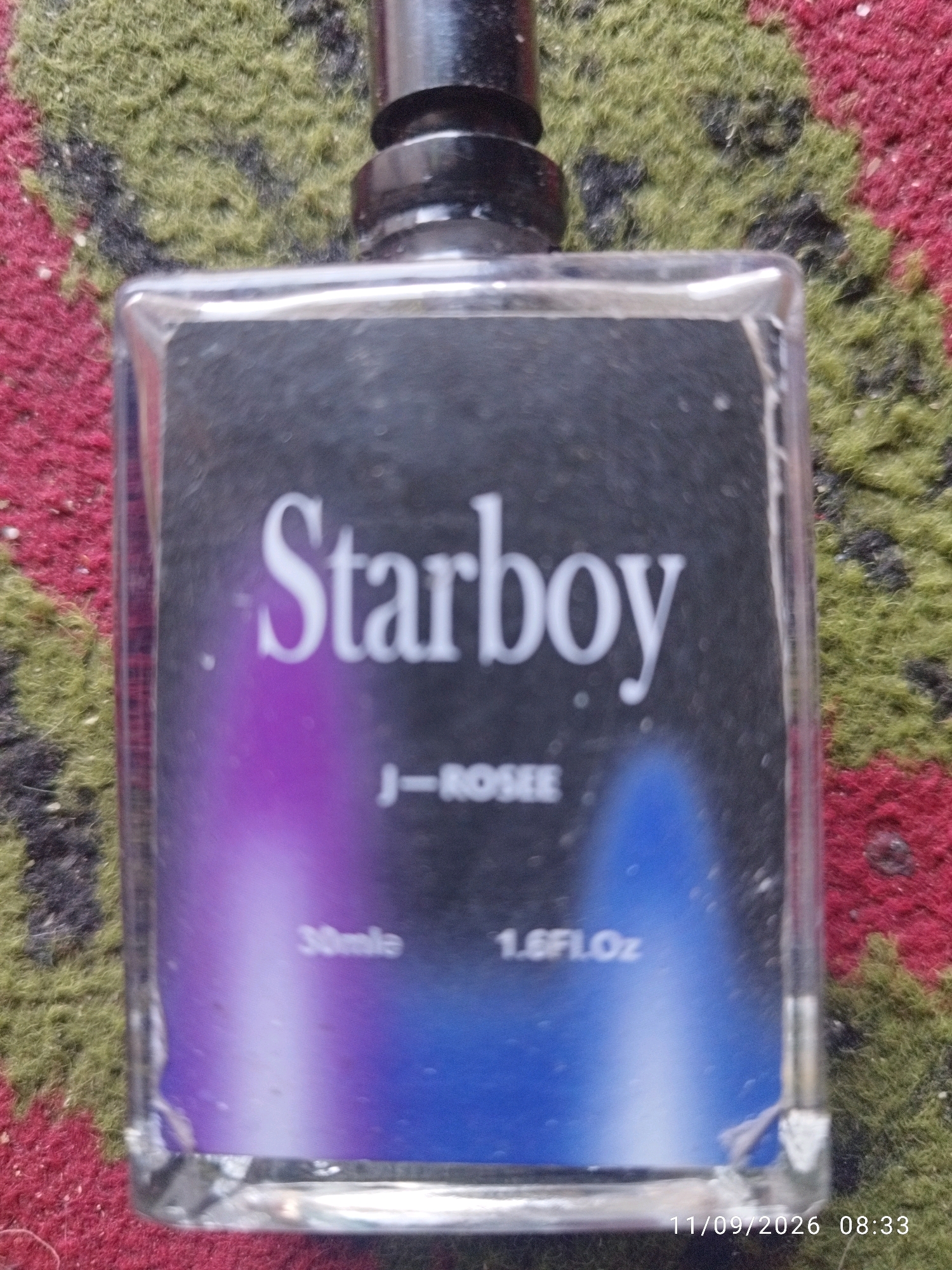 A clear, close-up, eye-level, indoor, overhead shot shows a clear glass perfume bottle with a black cap and a label featuring the word "Starboy" in large white serif font. Below "Starboy" is smaller white text that reads "J-ROSEE". The label has a gradient of purple and blue at the bottom. Towards the bottom left of the label, it says "30mle" and to the right, "1.6FLOz". The bottle is placed on a textured surface that appears to be a rug with red and green fibers. A timestamp "11/09/2026 08:33" is visible in the bottom right corner. The image does not show any people or activity specific to Mojokerto, Indonesia.