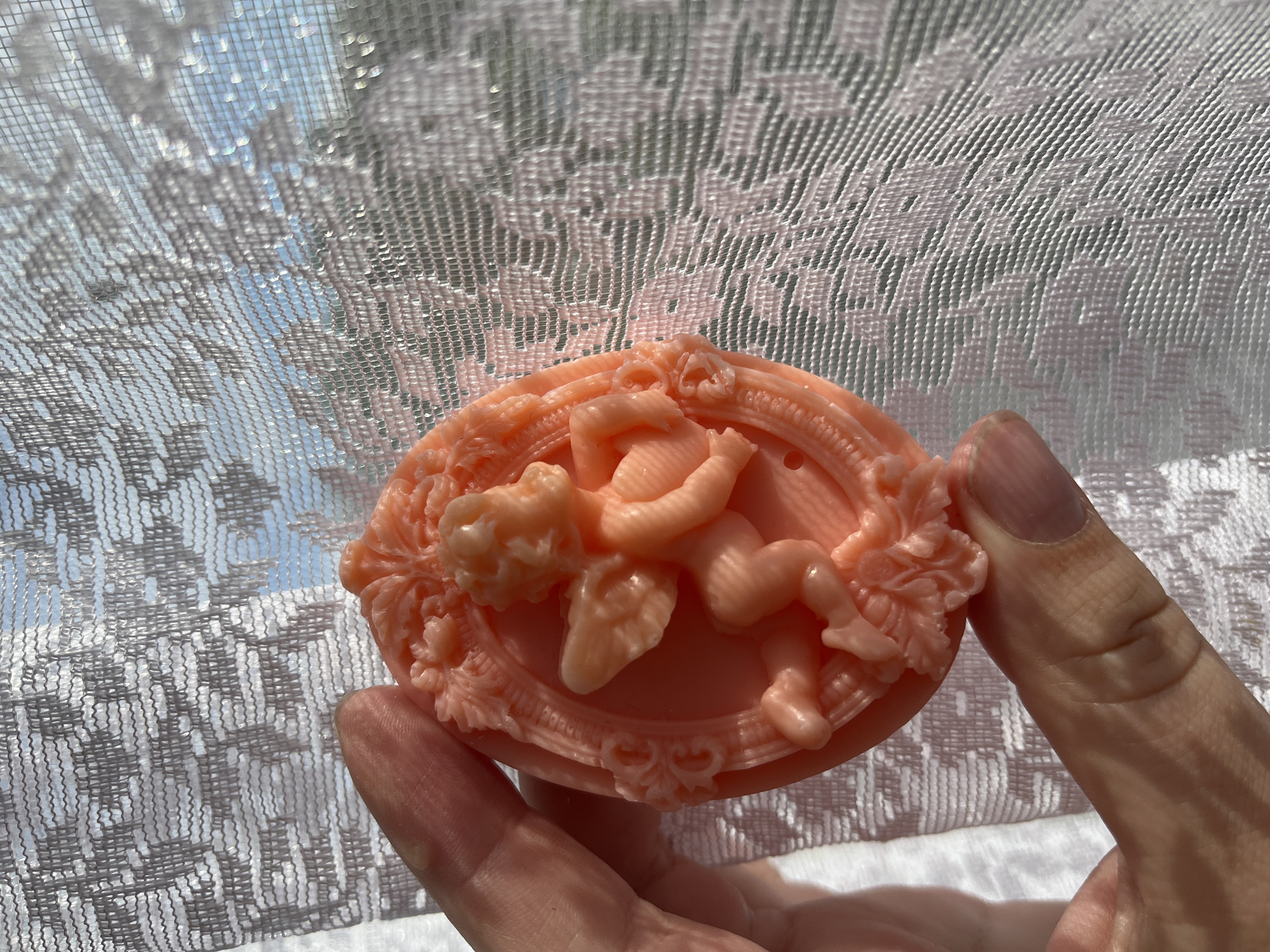 A person's hand holds a peach-colored oval soap with an ornate cherub design. The soap is placed in front of a sheer white lace curtain, which is illuminated by bright sunlight. The lace has a repeating geometric pattern. Through the curtain, the blue sky and some greenery are visible, suggesting it is daytime. The sunlight creates a dappled effect on the curtain. The overall impression is one of delicate beauty and natural light.