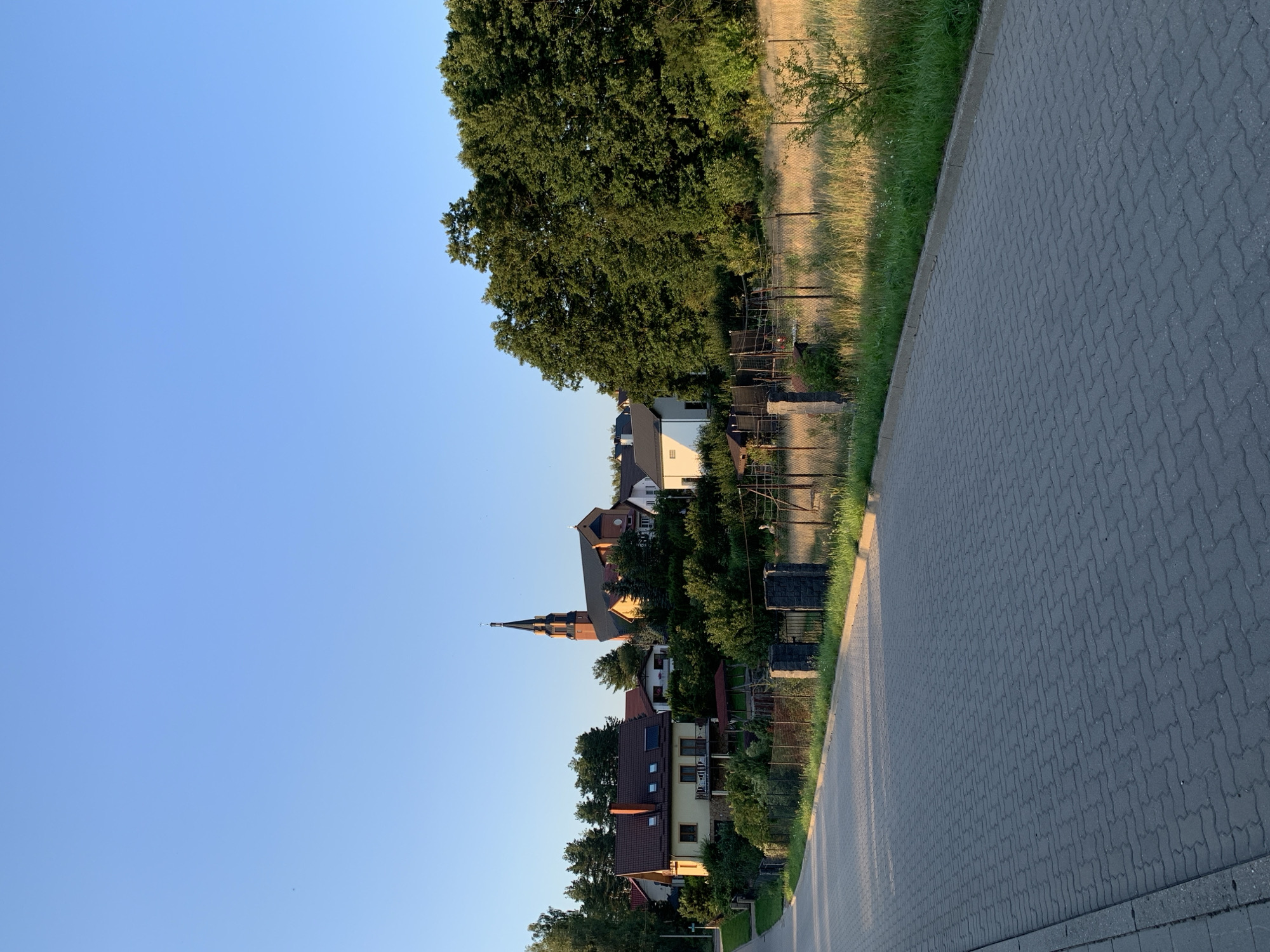 A paved road curves through a suburban area, leading towards a cluster of houses and a church spire under a clear blue sky. The road, constructed of grey interlocking pavers, occupies the foreground and extends into the midground, with a narrow strip of green grass and a wire fence bordering its right side. Beyond the fence, a field of dry, golden grass is illuminated by sunlight, contrasting with the lush green foliage of trees and bushes.

In the midground, several residential buildings are visible, characterized by red-tiled roofs and beige or white facades. One house in the left midground features multiple dormer windows. Dominating the right side of the midground and extending into the background is a dense stand of mature green trees, their leaves catching the sunlight.

The most prominent structure is a church, identifiable by its tall, dark-colored spire with a cross at its apex, and its red-brick building with a darker roof. The church is situated among the trees and houses, suggesting it is part of the local community. The scene appears to be a quiet, residential neighborhood, possibly in the late afternoon or early evening, given the warm light. No people are visible in the image. The setting evokes a sense of calm and rural tranquility within a developed area.