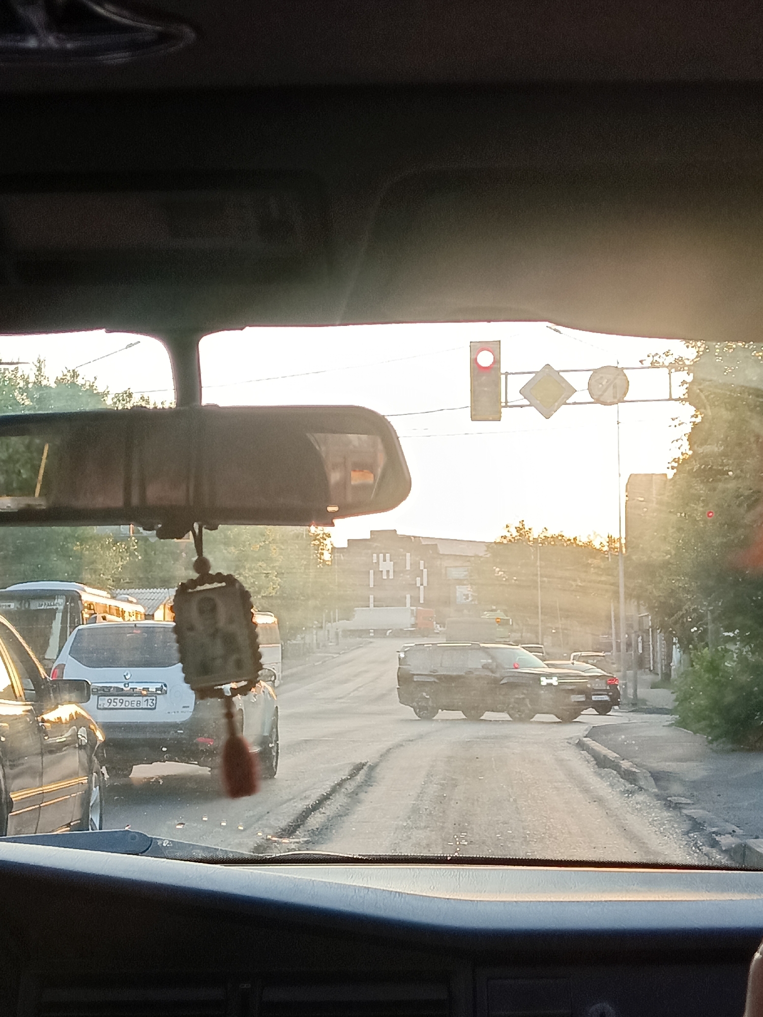 The image depicts a view from inside a vehicle, looking forward through the windshield. The scene is a street in Shymkent, Kazakhstan, bathed in the warm light of what appears to be late afternoon or early morning sun, causing lens flare and a bright sky. A traffic light is visible, with the red signal illuminated. Beside it are two road signs: a diamond-shaped yellow sign with a black border, and a circular sign with a red border and a diagonal red bar. In the middle ground, several cars are on the road, indicating traffic congestion. A white SUV with a license plate reading "959DEB 13" is prominent in the center. To the left of the SUV, a black vehicle is partially visible. Further ahead, a dark-colored SUV is also in traffic. The road surface appears to be asphalt and is somewhat uneven. Buildings line the right side of the street, and trees with green foliage are visible on both sides. Hanging from the rearview mirror inside the car is an ornament, possibly a religious icon. The interior of the car, including the dashboard and parts of the windshield frame, are visible in the foreground, suggesting the image was taken by a passenger or driver.