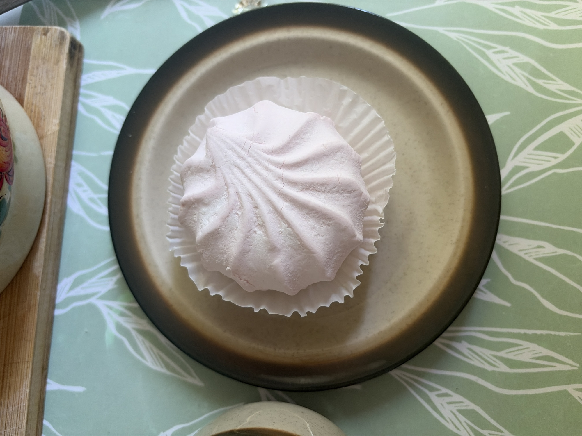 A single pink marshmallow sits in a fluted white paper liner on a brown-rimmed plate. The plate rests on a green placemat with a white leaf pattern. The scene is viewed from directly above.