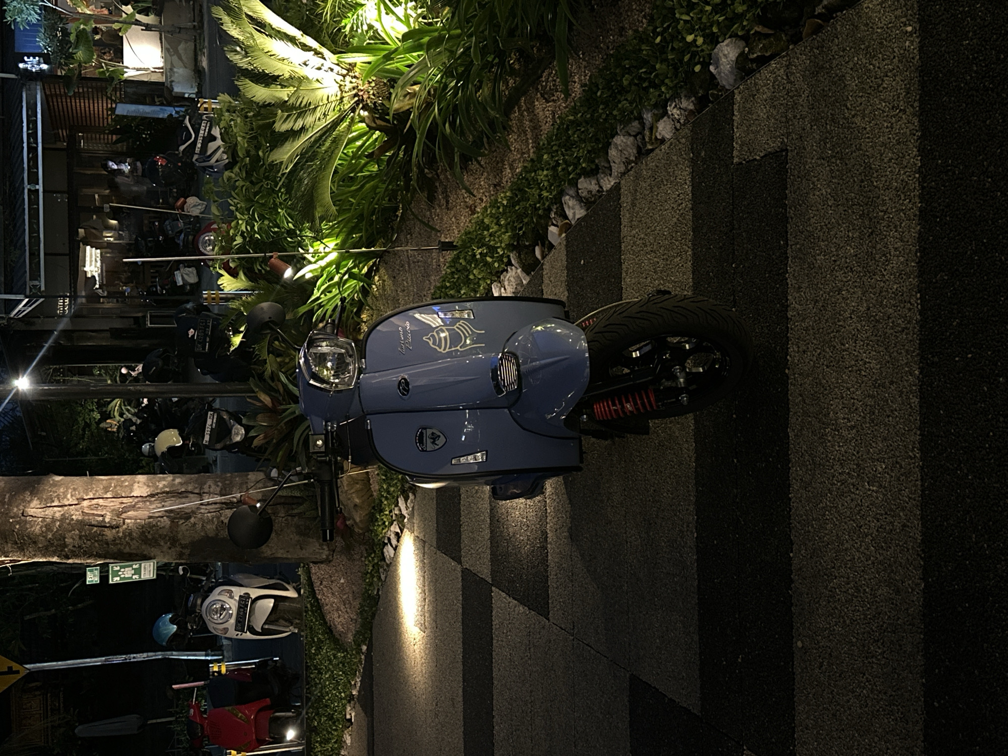 The image captures an evening scene in Mengwi, Indonesia, depicting a row of parked scooters outside a commercial establishment, likely a restaurant or cafe, illuminated by artificial lights. The time of day is clearly night, as evidenced by the dark surroundings and bright spotlights.
In the foreground, a prominent light blue custom scooter is parked on a patterned paved ground, featuring alternating dark and light rectangular sections. The scooter has "Mariano Piccolo" written on its front cowl, accompanied by a shell graphic. Its rear wheel and a red suspension component are visible. To the left, lush green vegetation, including a large palm tree, is dramatically lit from below by spotlights, creating a vibrant contrast with the darkness.
Further into the background, several other scooters and motorbikes are parked. These include a white scooter with a blue helmet beside it, partially obscured by a large tree trunk, and a red scooter visible behind it. Faint figures of people can be seen near the buildings, suggesting patrons or staff. Architectural elements of the buildings, including warm interior lights and slatted facades, are dimly discernible.
Visible text from license plates includes "DK 2768 FC1", "H 3174 MK", "DK 4209 FCT", and "DK 3391 CAB". A green "EXIT" sign is visible on the left, and Japanese text "アンリ" (Anri) is also present, possibly indicating a business name. The overall atmosphere is calm and relaxed, characteristic of an evening urban or resort setting.