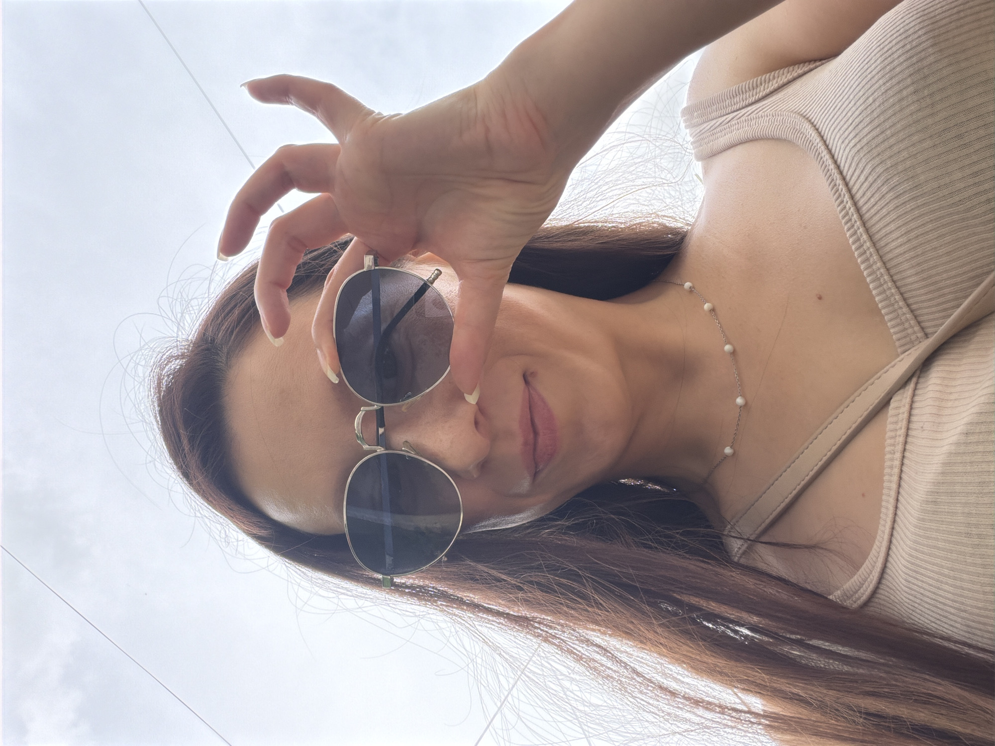 A young woman with long brown hair, wearing a beige ribbed tank top and a delicate pearl necklace, is pictured outdoors against a bright, overcast sky. She is looking towards the camera, and her right hand is raised, with her fingers obscuring part of her face and holding her sunglasses. The sunglasses have circular, dark lenses and a thin silver frame. A light-colored purse strap is visible across her shoulder. The background consists of a pale sky with some faint cloud formations and what appear to be electrical wires. The overall impression is a candid, casual portrait taken during the daytime. There is no information to suggest the location is in Tujmazy, Russia.