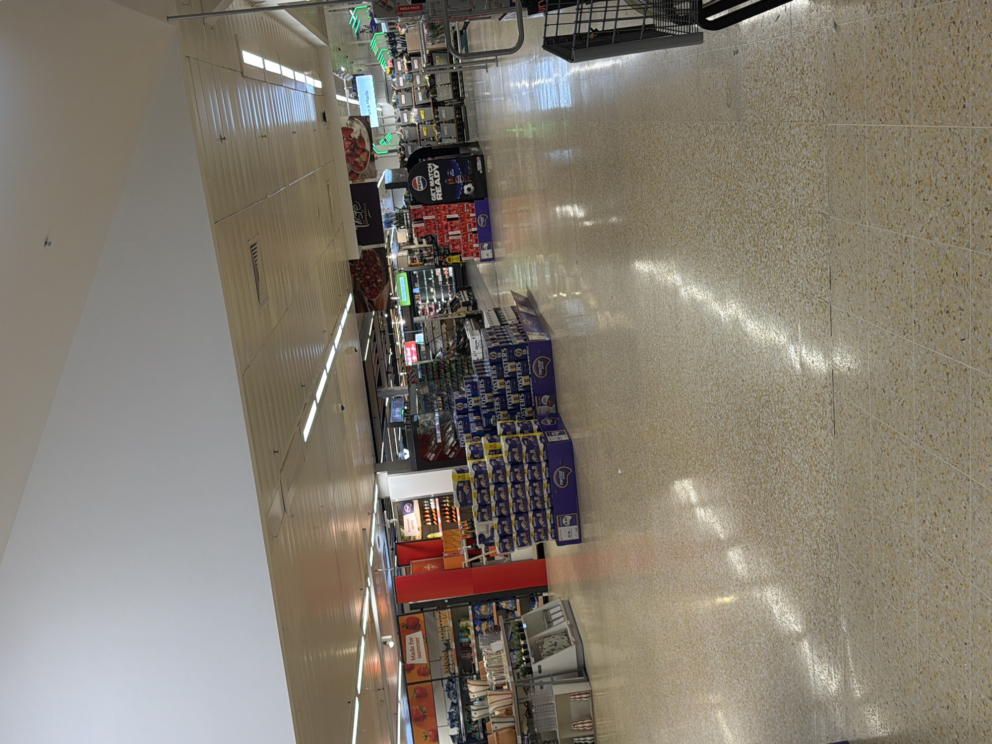 This is an interior shot of a supermarket aisle in Pity Me, United Kingdom. The aisle is long and well-lit with overhead fluorescent lights. The floor is a textured beige tile. Merchandise is arranged on shelves and in displays along both sides of the aisle. Brands visible include Pepsi, Fosters, and Nectar. The aisle also has signage related to 'Made for summer' products. There is a shopping trolley at the very top of the aisle. The angle suggests the photo was taken while standing still near the beginning of the aisle.