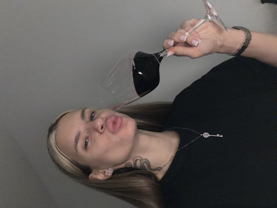 A fair-skinned woman with blonde highlights poses playfully with pursed lips, holding a wine glass filled with red wine. She's wearing a black shirt, a delicate key pendant necklace, and a wristwatch. A prominent snake tattoo adorns her neck. Her hair is long and straight, and she wears small, round earrings. The background is a plain gray wall.