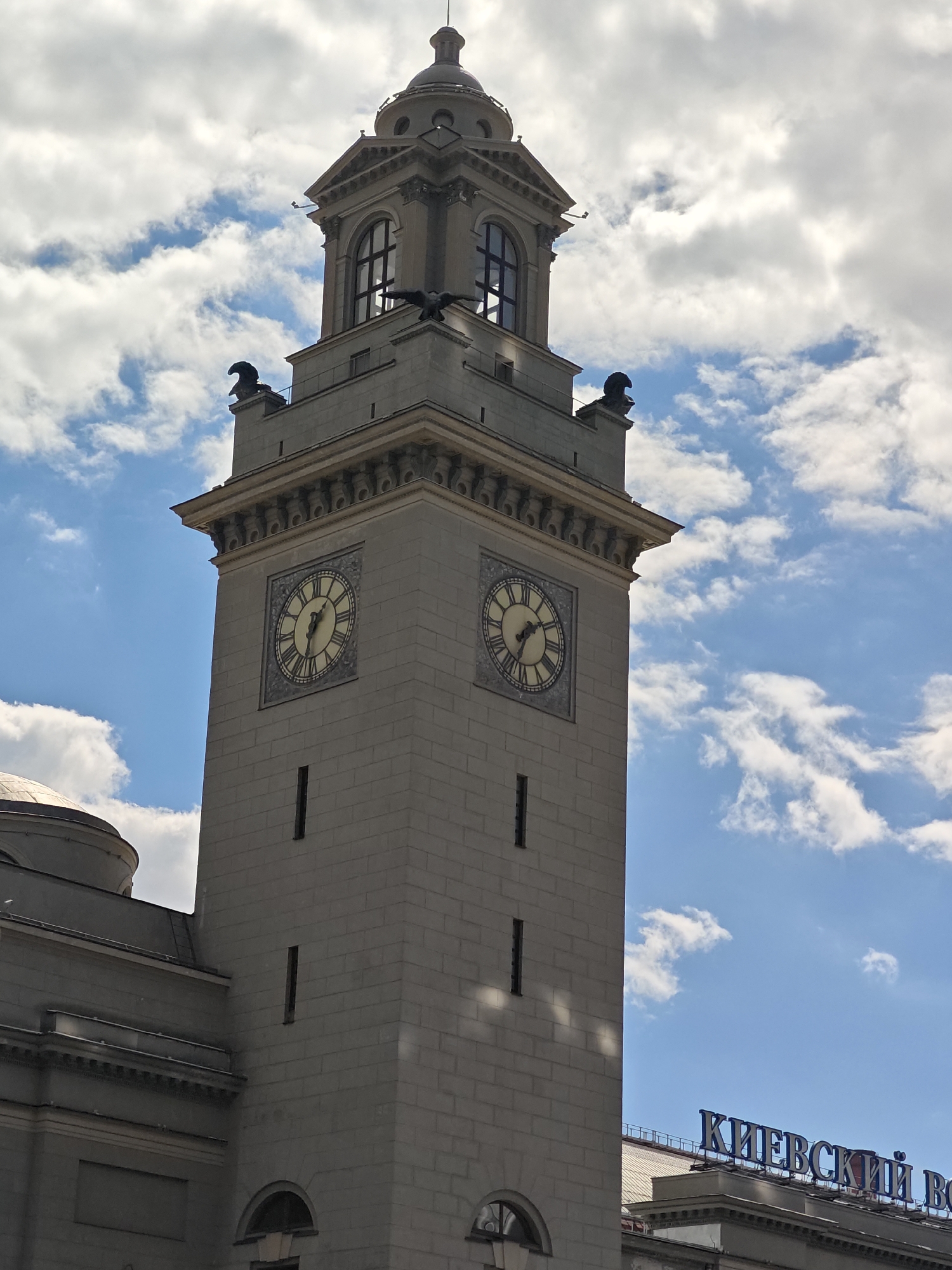 This media file features a close-up, low-angle shot of a grand, stone clock tower against a bright blue sky filled with fluffy white clouds. The tower has a classical architectural style, with decorative cornices and arched windows. Two large, circular clocks with Roman numerals are prominently displayed on the tower's facade, one on each visible side. The clock hands on the left face appear to show approximately 10:07, and on the right face, approximately 10:09. Perched atop the tower's corners are dark, sculpted figures that resemble eagles.
To the left of the tower, a curved, grey dome and part of a building with classical detailing are visible. To the right, in the lower portion of the image, blue lettering spells out "КИЕВСКИЙ ВОКЗАЛ" (KIEVSKY VOZCAL) in large, stylized font, indicating the location as Kievsky Railway Station in Moscow, Russia. The lighting suggests it is daytime, likely late morning or early afternoon, with clear and pleasant weather. The image evokes a sense of grandeur and historical significance, focusing on the architectural marvel of the station's tower. No people are visible in the frame.