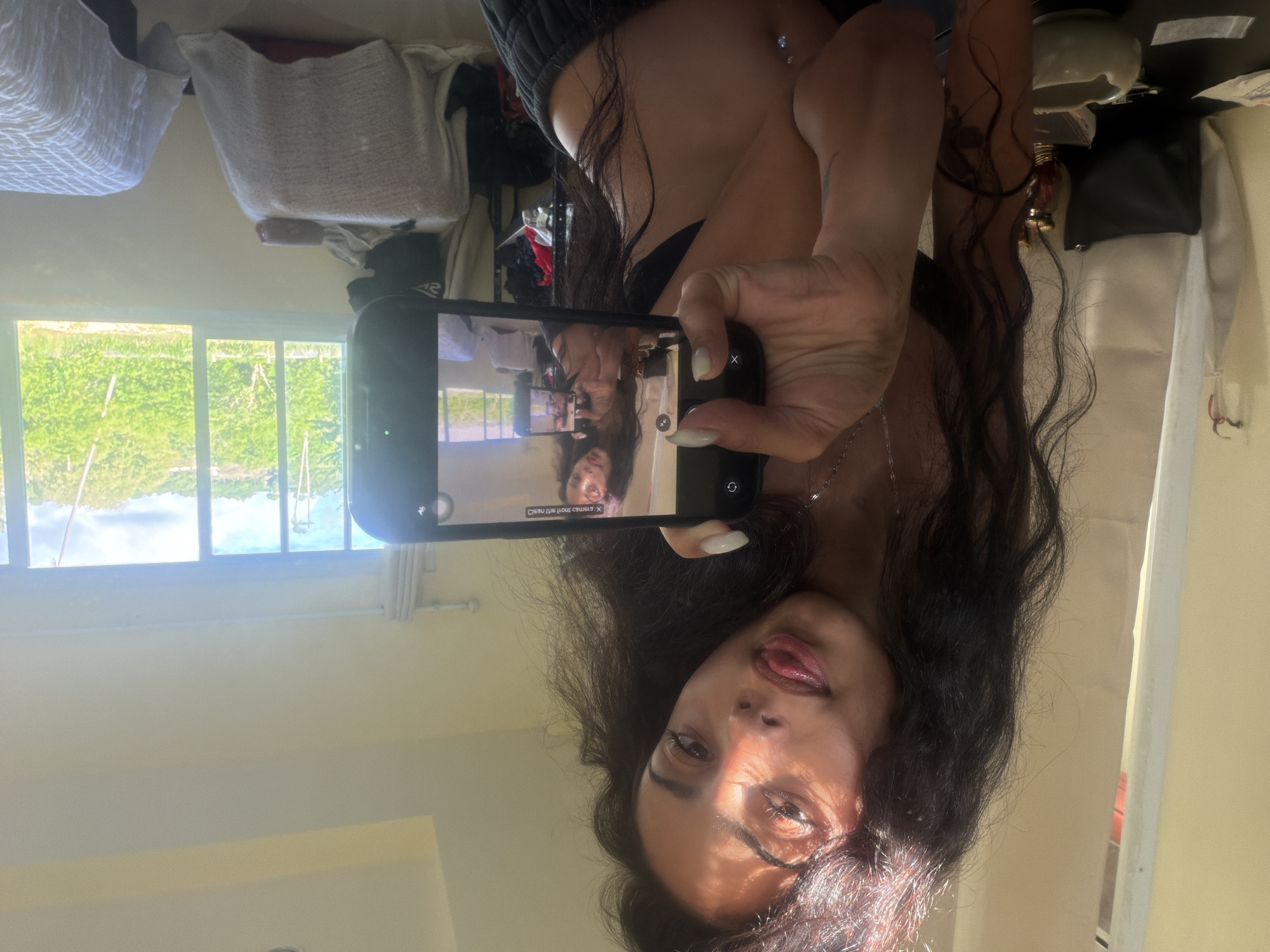 A young woman with long, dark, wavy hair is taking a selfie with her smartphone. She is indoors, near a window that offers a view of lush green trees and a bright sky, indicative of a tropical location such as Baan Bo Phut, Thailand. The room appears to be a bedroom, with what looks like a bed covered with a white quilt on the right side of the frame. The woman is wearing dark clothing, and a necklace is visible around her neck. Her tongue is sticking out slightly as she poses for the picture. The phone's screen shows a reflection of the woman and the room. The overall lighting suggests it is daytime.