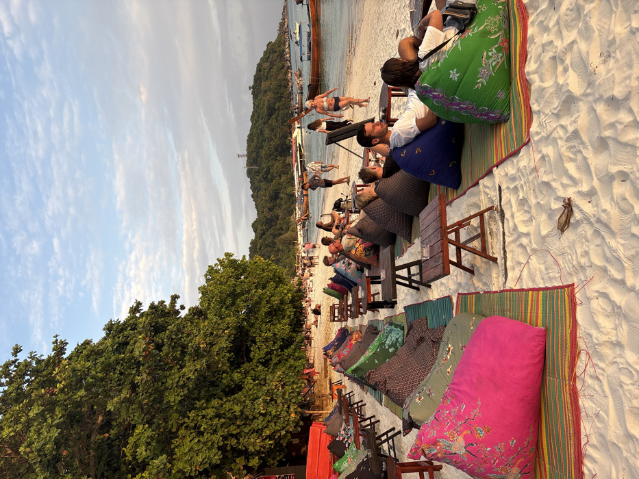 The image captures a beach scene at sunset or golden hour in Ban Ko Li Pe, Thailand. The sky exhibits a gradient of light blue with scattered, wispy white clouds, illuminated by warm, low-angle sunlight.
In the foreground, a white sandy beach hosts multiple rows of beach seating arrangements. Each setup consists of a brightly colored, patterned beanbag chair placed on a striped woven mat, accompanied by a small, dark wooden table. Beanbag colors include pink, green, blue, and various multi-patterned designs, while mats feature green, red, and yellow stripes. Several tables contain small ashtrays.
Multiple individuals are visible, primarily seated or reclining on the beanbags. In the immediate foreground, a person is seated on a bright green beanbag, and another on a blue beanbag. Further back, a man in a white t-shirt is seated on a blue beanbag. Other patrons are scattered among the seating, some conversing, others facing towards the water.
Along the sandy shoreline, numerous people are walking or standing. A woman in a bikini top and shorts is seen walking towards the right, and a man in shorts stands near the water's edge. Additional groups of individuals populate the distant beach area.
The calm, light blue-green sea in the mid-ground contains several traditional Thai longtail boats and smaller motorboats, either anchored or moored. To the far left, a dense canopy of large green trees borders the beach. In the background, a heavily forested island or hill rises from the water, topped by a distinct communication tower or antenna structure.