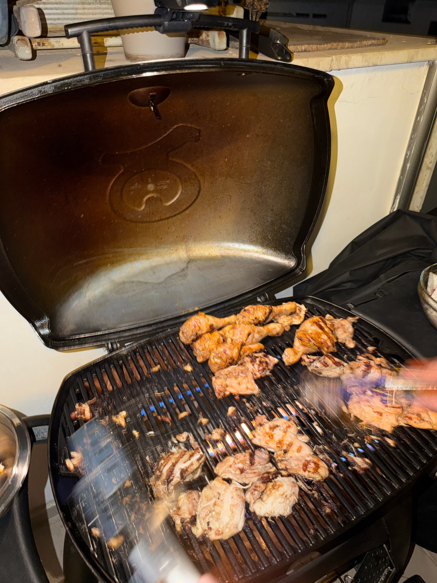 An open grill with chicken pieces is pictured in what appears to be an outdoor urban setting in Tel Aviv, Israel, with a residential building wall visible in the background. The grill, a Weber model, features a dark lid, slightly ajar, revealing a circular logo etched into its interior. The grates are laden with chicken drumsticks and thighs, some displaying char marks from the cooking process. Blue flames are visible beneath the chicken, indicating the grill is active. A metallic tool, possibly a spatula, is being used to turn the chicken, creating a blurred effect due to motion. To the right, a black cloth cover for the grill is draped, and a glass bowl containing food is partially visible. The overall scene suggests a domestic outdoor cooking event.
