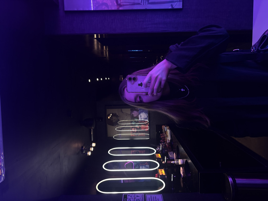 A female subject is depicted taking a mirror selfie in a dimly lit public restroom or lounge area within Mexico City, Mexico. The environment features dark walls and ceiling, illuminated by purple ambient lighting and white neon-bordered oval mirrors positioned above a long, dark countertop. The subject, occupying the right half of the frame, holds a light-colored smartphone, which partially obscures her face. She has long, light brown hair and wears a dark, long-sleeved top with a visible cross pendant necklace. Her left hand, holding the phone, displays light-colored nail polish. On the counter, multiple small white dishes containing small white objects, a pump dispenser, and folded items are visible. A large framed poster featuring multiple male figures, prominently including Jim Morrison, is mounted on the back wall. To the far left, a white sign with black pictograms and Spanish text is affixed to a dark wall, above a silver-toned trash receptacle. The ceiling contains recessed lights. A textured purple wall element is visible on the far right.