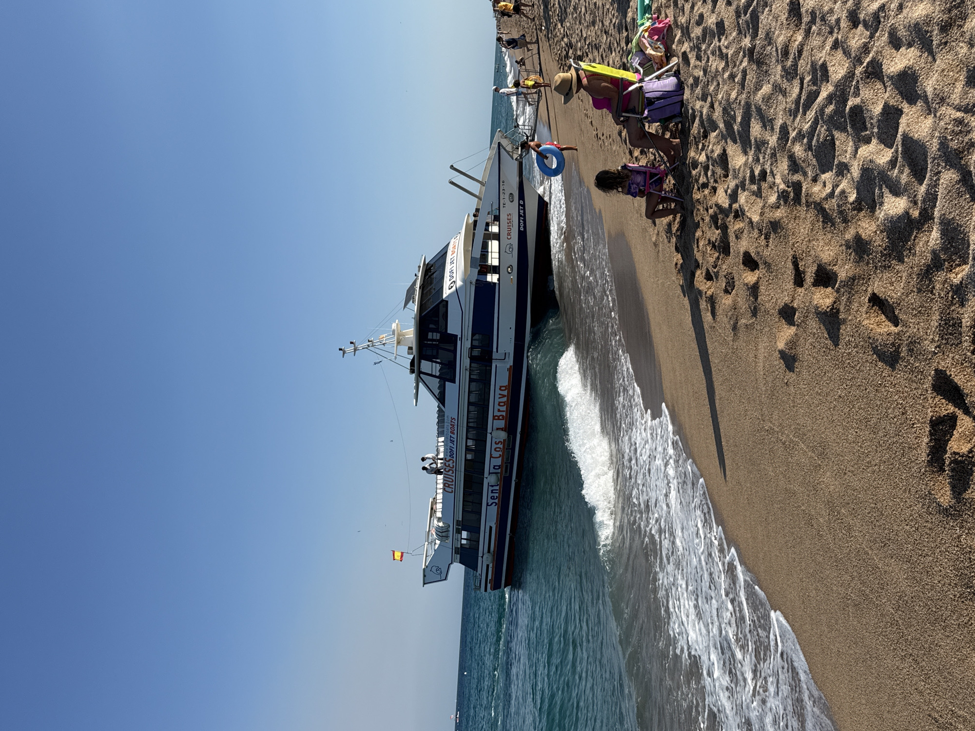 In Blanes, Spain, a large, white and blue passenger boat named "Sent La Costa Brava" is approaching a sandy beach. The boat has "CRUISES" and "DOFI JET BOATS" written on its side. Two people are visible on the upper deck of the boat. The sea is calm with gentle waves lapping at the shore. Several people are on the beach, including a woman in a pink swimsuit sitting in a yellow chair, and a young girl in a purple swimsuit sitting in a pink chair. A young boy holding a blue inner tube stands near the water's edge. The beach is composed of fine sand with footprints visible. The sky is clear and blue.