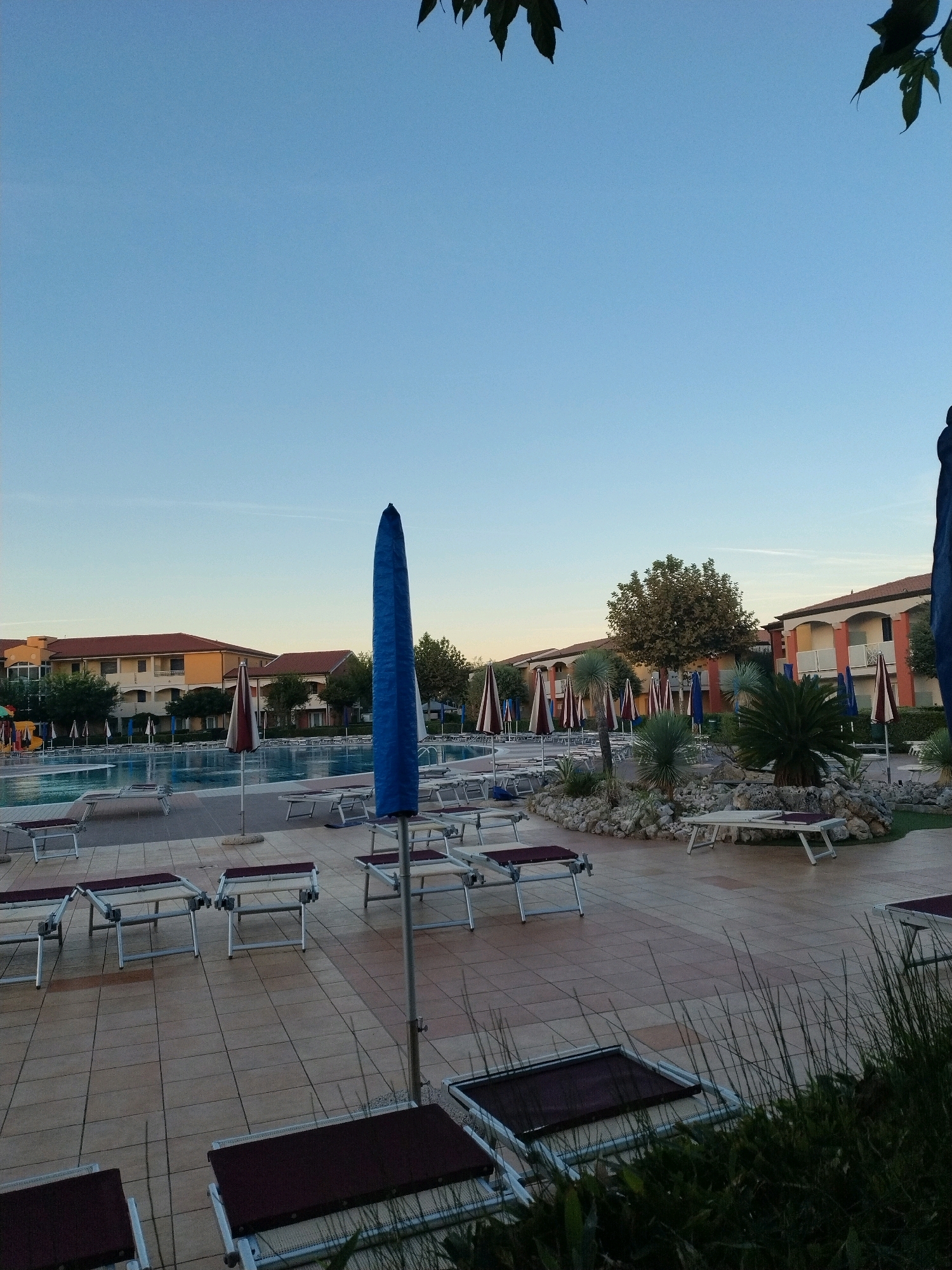 The image depicts an outdoor swimming pool area at a resort in Caorle, Italy, likely during the early morning or late evening due to the soft, clear sky and lack of direct sunlight. The foreground features several empty lounge chairs, some with dark cushions. A tall, folded blue umbrella stands prominently in the mid-ground. In the background, a large swimming pool reflects the pale blue sky. Beyond the pool, a row of buildings with orange and cream facades and red-tiled roofs are visible, suggesting accommodation units. Several smaller, striped umbrellas are positioned around the pool area. Lush green trees and shrubs, including palm-like plants, are interspersed throughout the scene, adding a tropical feel to the resort. The ground is paved with tan tiles. There are no people visible in the image, creating a serene and unoccupied atmosphere.