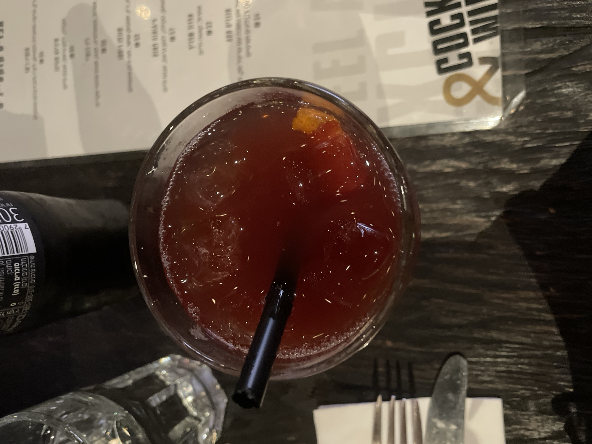 The image shows a bird's-eye view of a red cocktail in a glass, served with a black straw, likely in a restaurant or bar setting. The menu is partially visible behind the drink, featuring the words "COCKTAIL & WINE." In the lower right corner, there's a fork and knife on a napkin. A bottle and another glass are also visible in the lower left corner. The text on the bottle is in Hebrew, suggesting the location is likely in Tel-Aviv, Israel.
