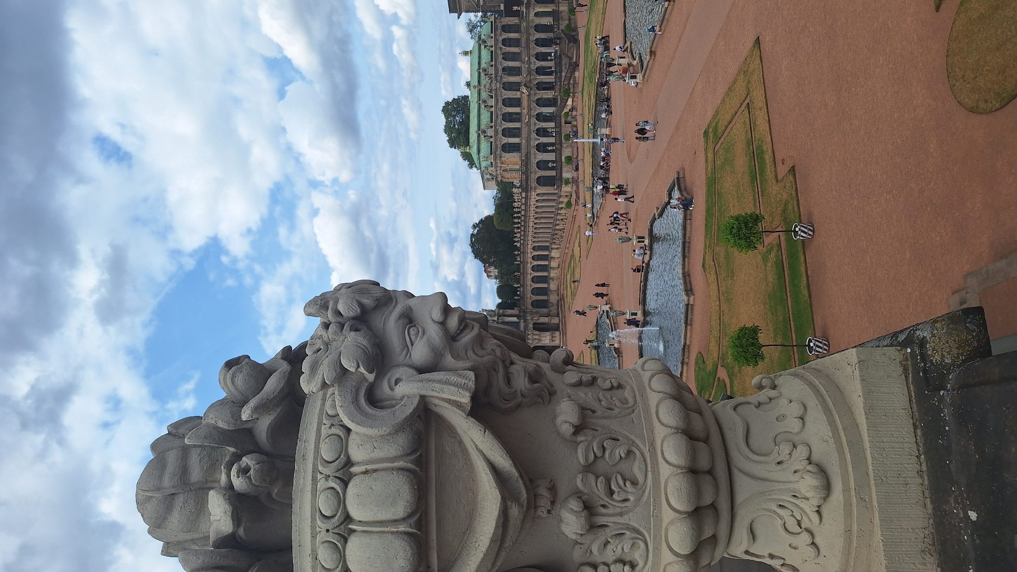 The image captures an outdoor scene at the Zwinger Palace in Dresden, Germany. In the foreground, a large, ornate stone urn with a carved grotesque face and decorative floral motifs dominates the left side of the frame. The urn is positioned on a stone ledge, with its base extending downwards. In the background, the grand architecture of the Zwinger is visible, characterized by its Baroque style, featuring long colonnades with arches and a prominent central building with a green roof.
The scene is a courtyard or garden area, paved with reddish-brown gravel. Several water fountains are active, with water jets rising from them. People are scattered throughout the courtyard, walking, sitting, and observing the surroundings. Two small, conical trees in striped planters are positioned on the gravel area. The sky above is partly cloudy, with patches of blue sky visible amidst fluffy white and gray clouds. The lighting suggests it is daytime. The perspective is from a slightly elevated viewpoint, looking down and across the courtyard towards the palace.