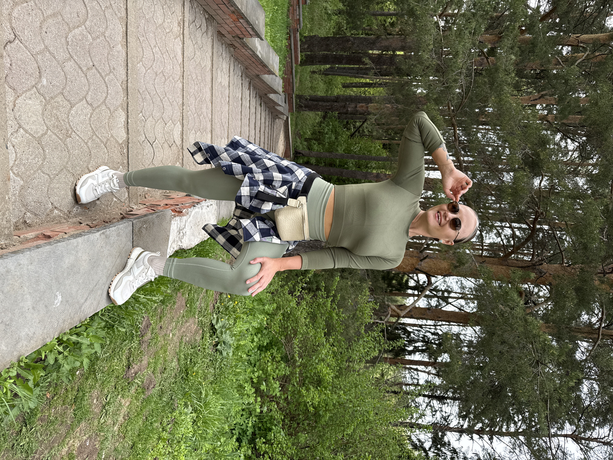 The image shows a woman in athletic wear posing outdoors in Chaikovsky, Russia. She is wearing a green long-sleeved crop top and leggings, white sneakers, and a black and white checkered shirt tied around her waist. Sunglasses are perched on her face. She is standing near a stone walkway with a brick border, adjacent to a grassy area with trees.