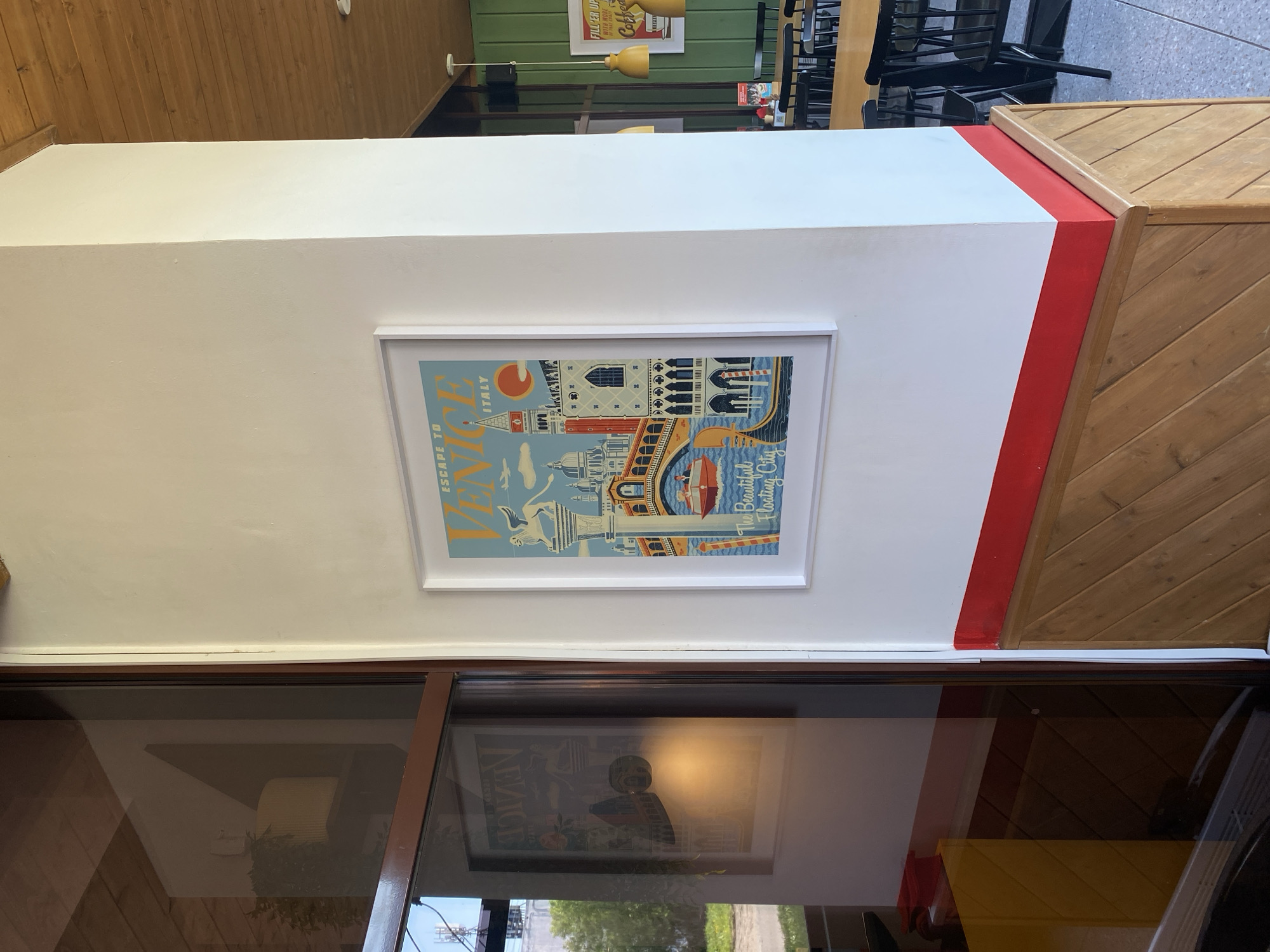 The image shows a white wall with a framed poster of Venice, Italy, hanging on it. The poster has a blue background and features illustrations of Venetian landmarks such as St. Mark's Basilica and gondolas. Above the wall is a wooden ceiling, and to the right, a wooden structure with a red trim. Below the wall is a window with a view of greenery outside. A reflection of another Venice poster can be seen in the window glass. The location is not Izhevsk, Russia.