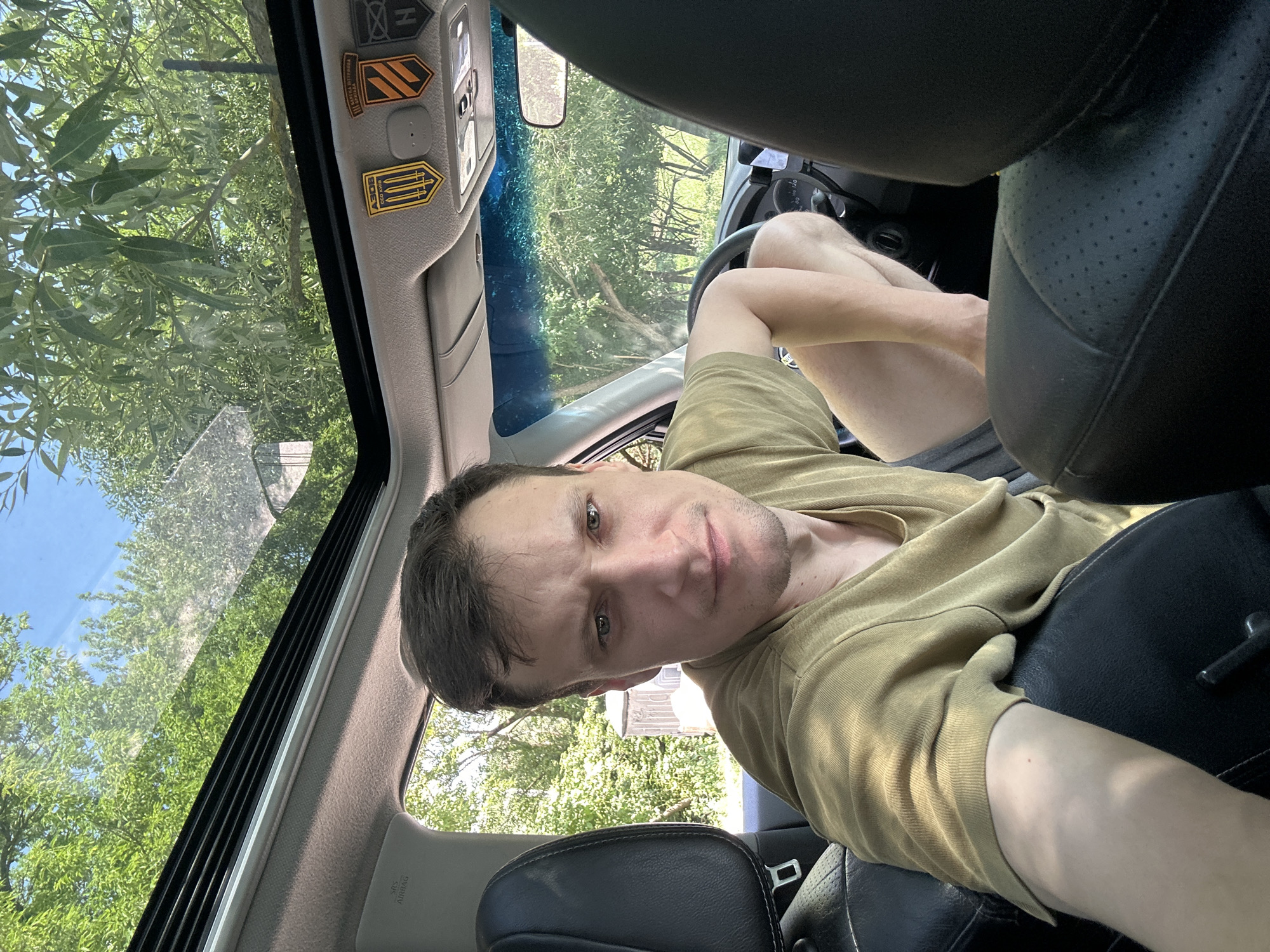 A man is taking a selfie in a car with an open sunroof on a sunny day. He is wearing a tan t-shirt and dark shorts, with his left arm resting on the seat. He is looking directly at the camera with a neutral expression. The car's interior is visible, including the dashboard, steering wheel, and seats. Outside the car, lush green trees and a blue sky are visible through the open sunroof. Two patches are affixed to the car's interior ceiling; one patch depicts three stylized swords on a yellow background and the words "АЗОВ СОО КИЇВ" in Ukrainian, the other is a black and orange chevron.