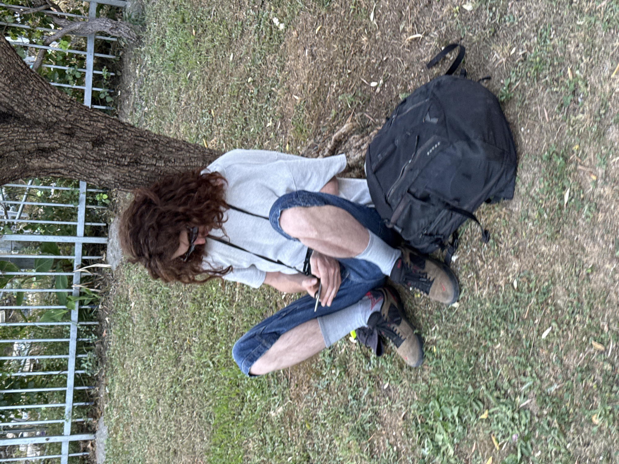 A person with long, curly brown hair and glasses is sitting on the ground next to a tree. They are wearing a light-colored long-sleeved shirt, blue jeans with a hole in one knee, light gray socks, and brown and black hiking shoes. The person is holding a cigarette in their right hand. A black backpack is lying on the ground behind them. The scene appears to be outdoors, with grass and dirt covering the ground. A metal fence is visible to the left of the frame, with some greenery behind it. The image is taken from a slightly elevated angle, looking down at the person. The lighting suggests it could be daytime, possibly late afternoon or early morning. The overall impression is one of relaxation or rest. There is no visible text in the image.