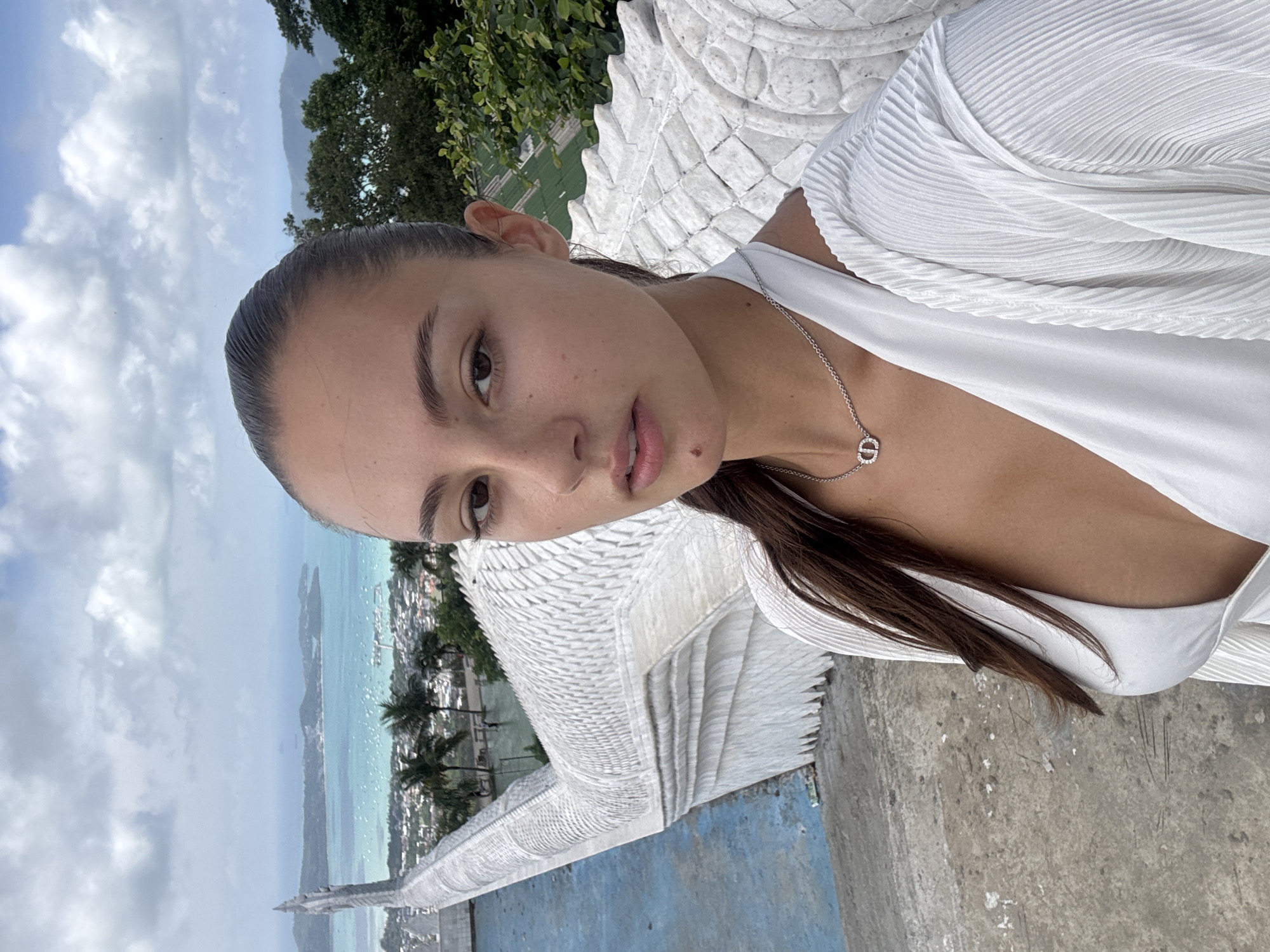 The image shows a woman in a white top and silver necklace, facing the camera at an angle. She is standing at what appears to be the Karon Viewpoint, in Karon, Thailand. The backdrop shows a scenic view of the coastline, including the ocean, beaches, and cityscape, with palm trees visible below. A white stone structure, likely part of the viewpoint's architectural design, is in the foreground beside her. The sky is overcast with clouds.