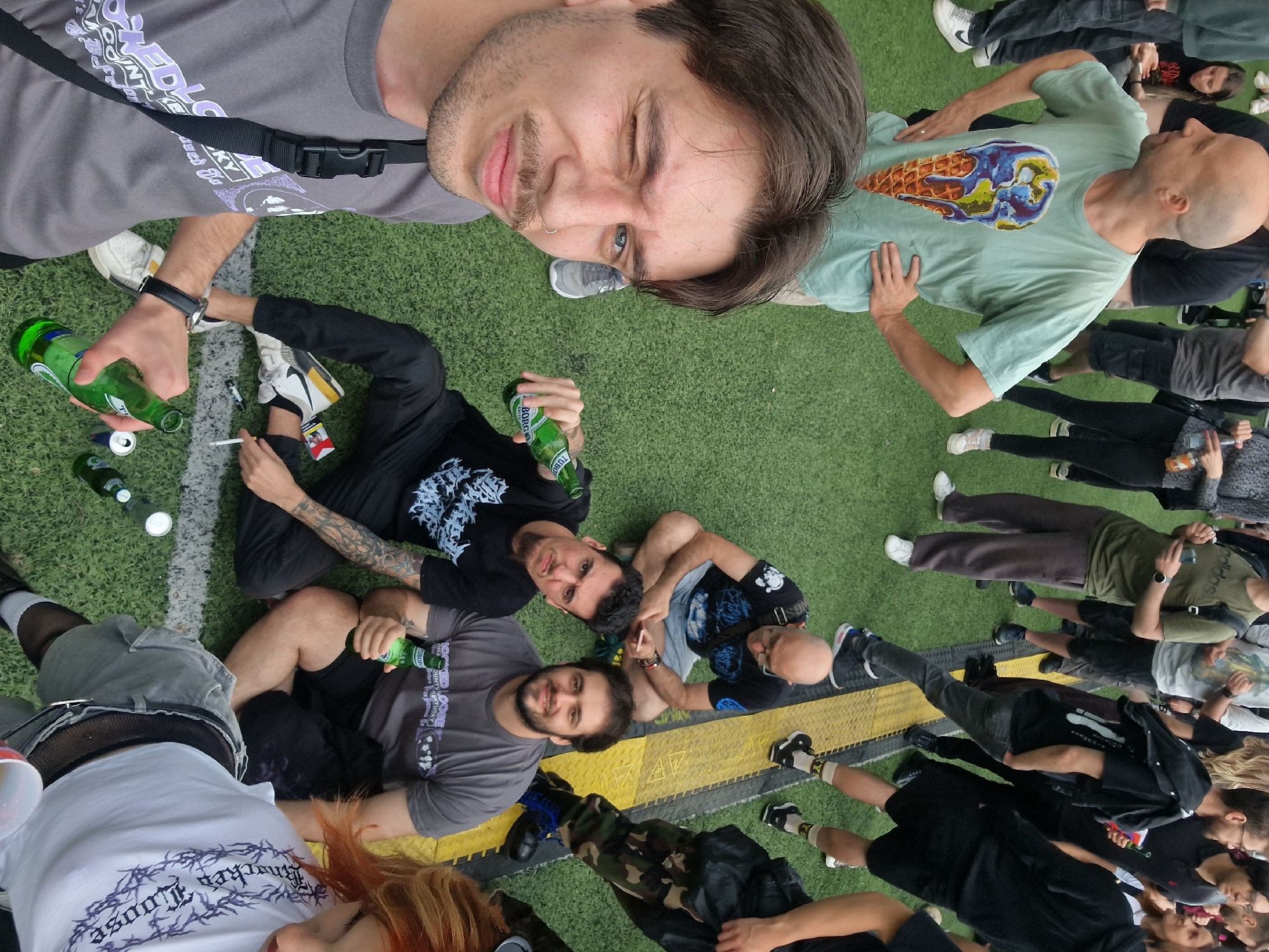 A group of approximately 20 people are gathered on a field with artificial turf and yellow painted lines. The scene is shot from a high angle, possibly from an elevated position or using a wide-angle lens pointed downwards. Many of the individuals are sitting or lying on the grass, and a few are holding beer bottles and cigarettes. There is a mix of clothing styles, ranging from casual T-shirts and shorts to more alternative looks with fishnet stockings and darker attire. The background is somewhat blurred due to the depth of field. The location in the city of Unknown, Unknown is unknown, because it is not recognizable from the image.