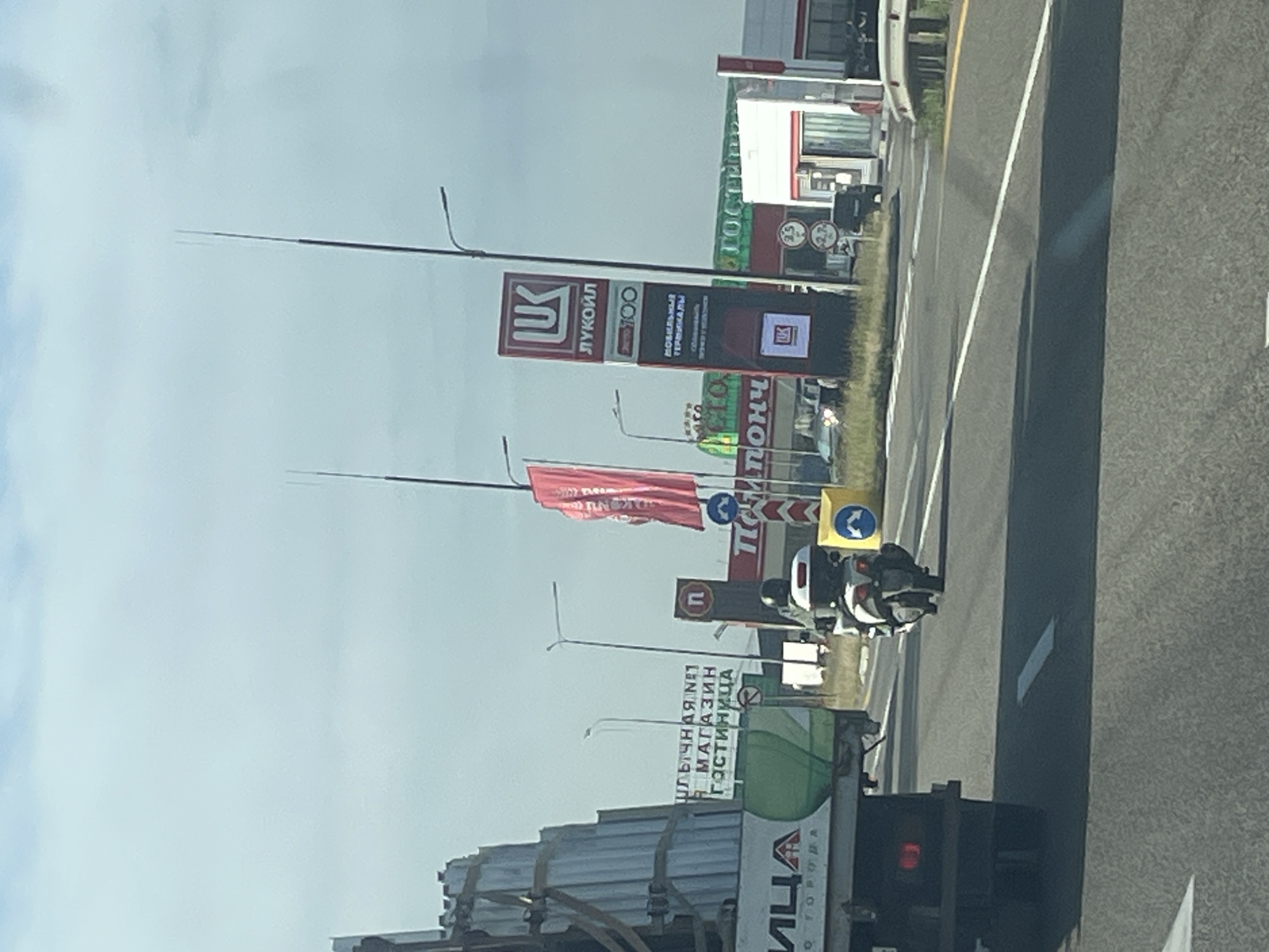 A motorcycle rider, wearing a helmet and white jacket, travels on a multi-lane paved road. The rider is on a white motorcycle with a black seat and a white storage box on the back. The road is flanked by a guardrail and a grassy shoulder. In the background, there are several buildings and signs. A large red sign with "ЛУКОЙЛ" (Lukoil) is visible, along with a gas station. A truck is parked on the left side of the road. The sky is overcast with clouds. The scene appears to be a highway or main road in or near the city of Tugurgoy, Russia, indicated by the Lukoil branding common in Russia.